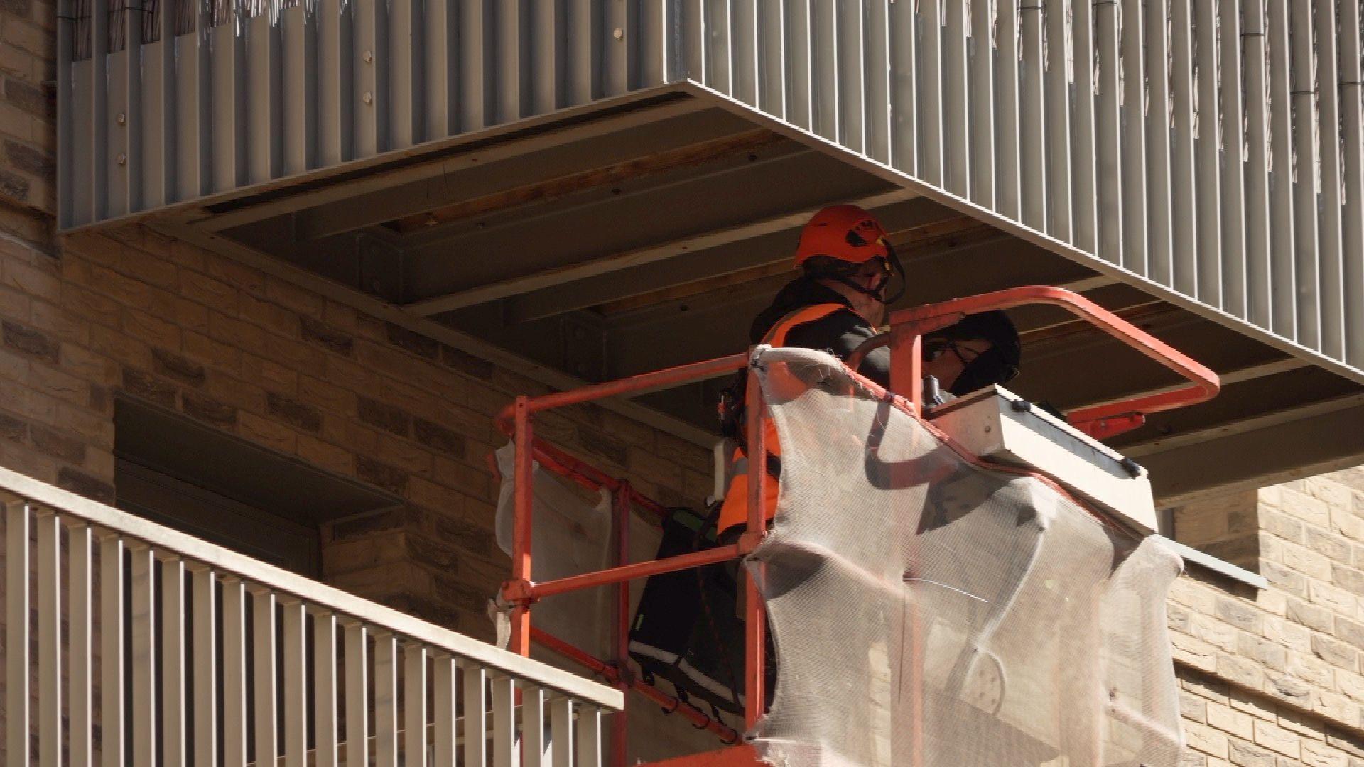 Two engineers in orange helmets and black uniforms inspect the underside of a balcony from a cherry picker