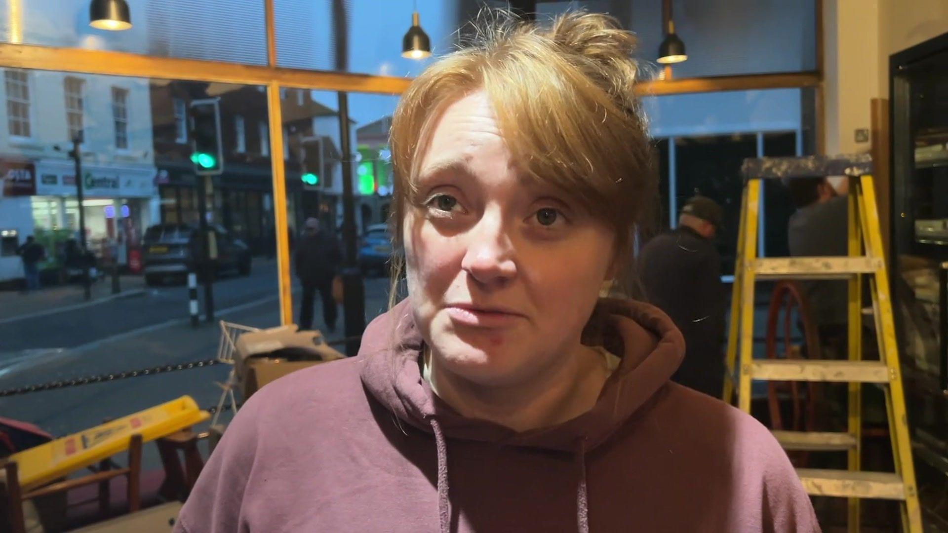 Sarah Redrup giving an interview at her bakery. She is wearing her hair in a pony tail and has a dark red hoody on. A handyman is working behind her. There is a yellow ladder inside.