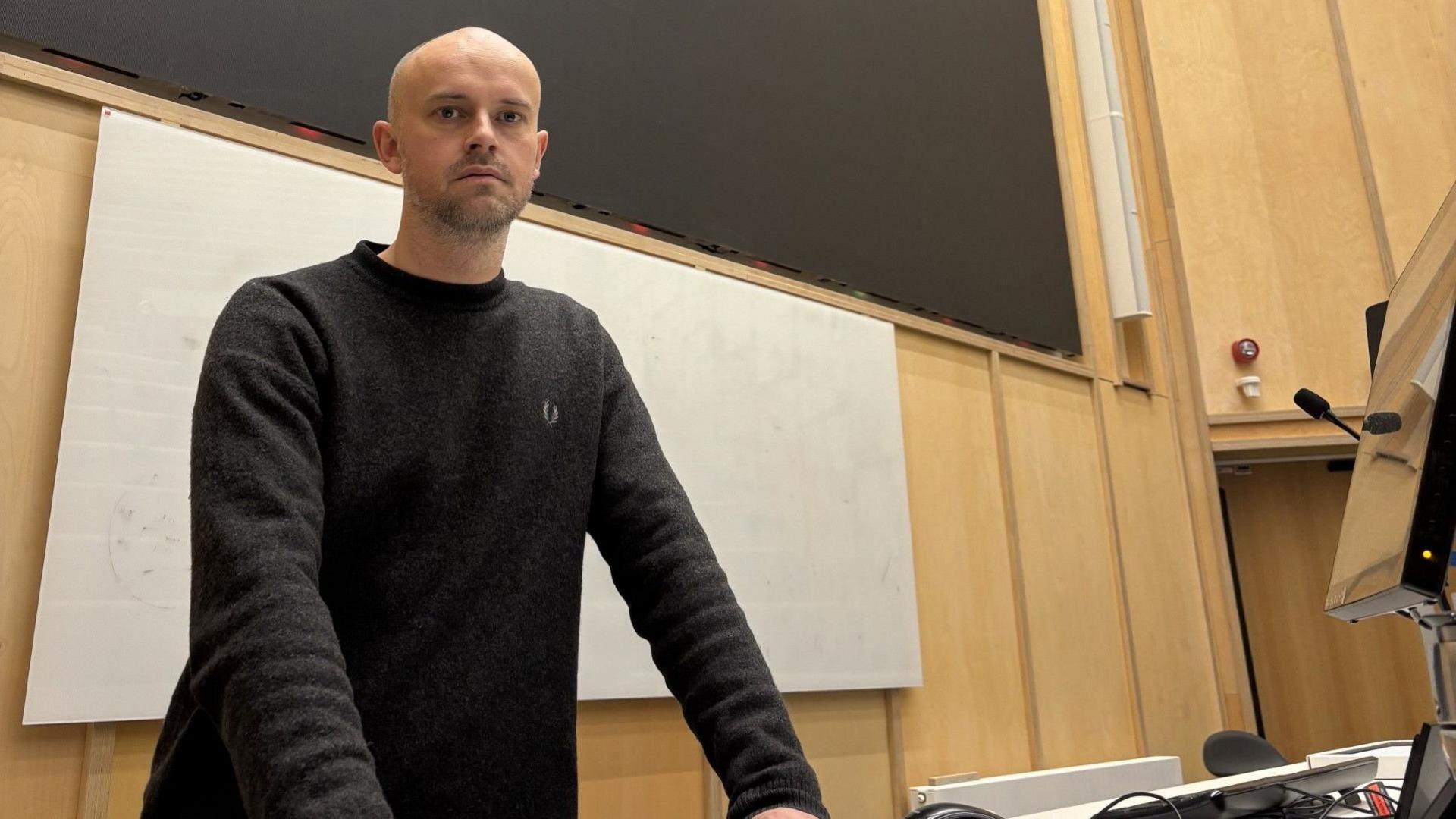 Dr Joey Whitfield standing in a lecture theatre. He is wearing a black jumper and looking at the camera.