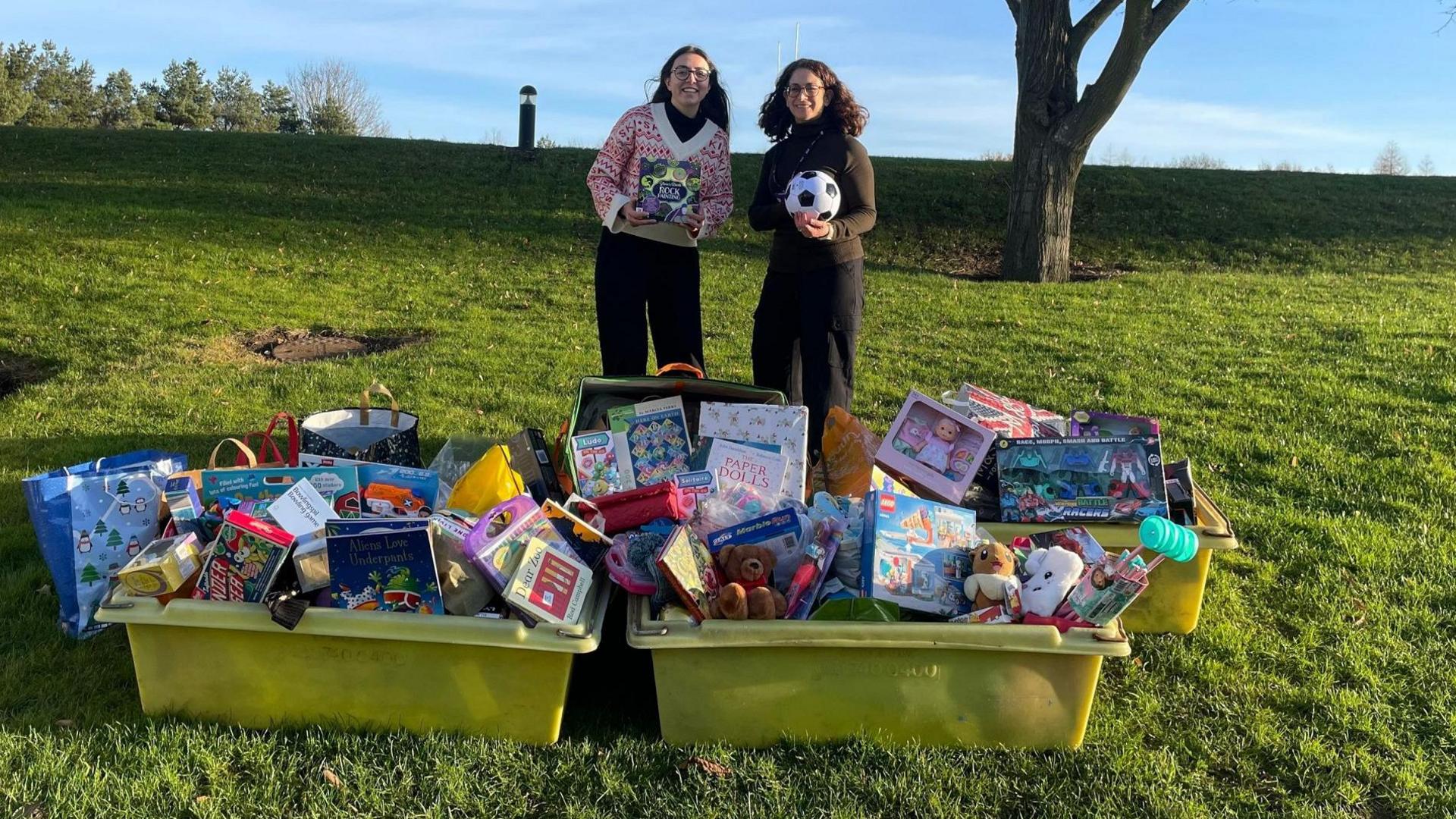 Two women - one in a Christmas jumper - stand behind two large, yellow plastic boxes filled with children's presents such as books, dolls and teddies. They are standing in a park.