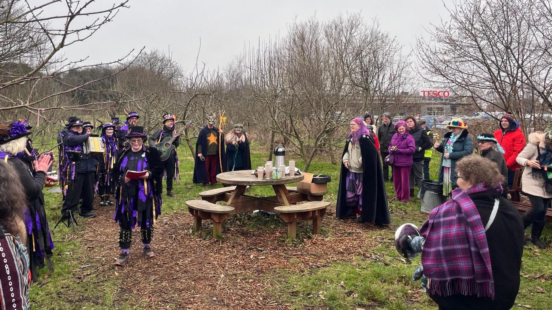 A group of people in brightly coloured clothers gather round a wooden circular table in the middle of an orchard. Bare trees stand on all sides.