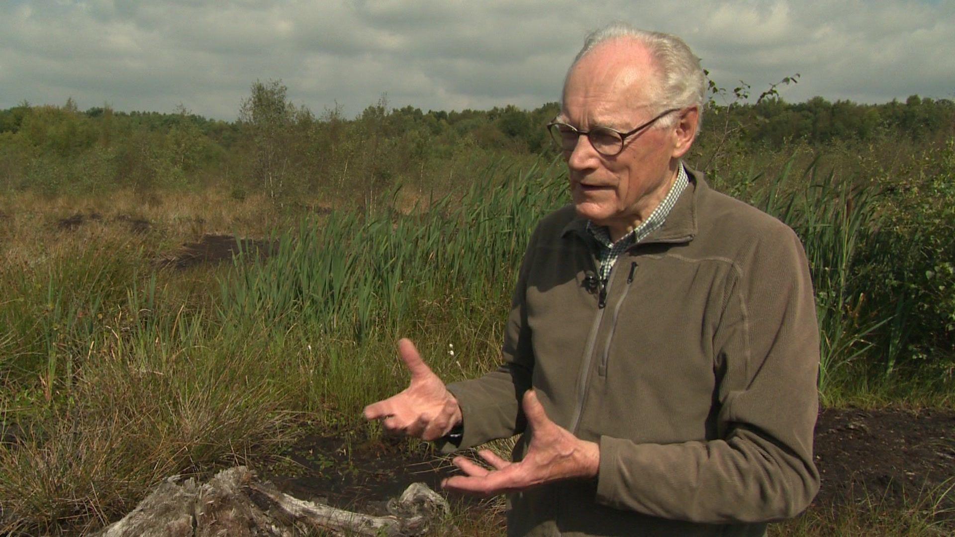 Professor John Handley, who wears glasses and wears a dark green fleece, standing before a grassy and overgrown area of Lindow Moss peat bog