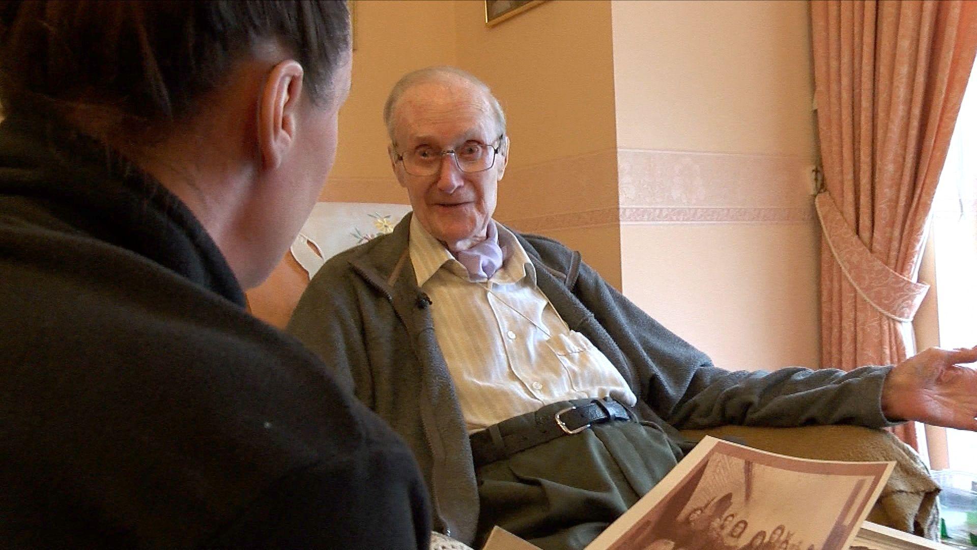 Ron Champion is in the background sitting in his armchair. He's wearing a pale shirt, grey cardigan and grey trousers. In the foreground his befriender, Sarah, has her back turned towards Ron and a sepia photograph in her hand.