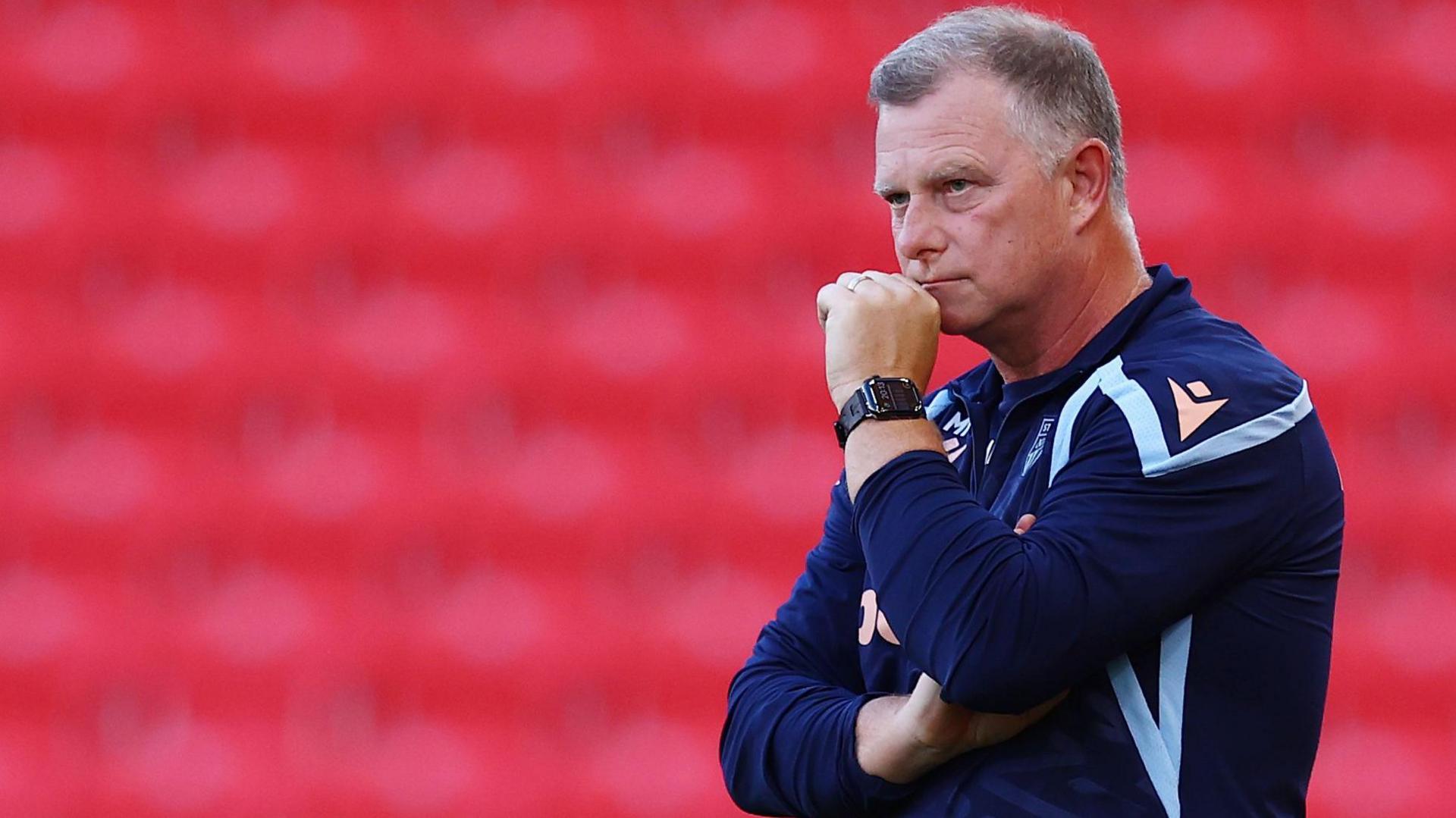 Stoke City manager Mark Robins watches from the touchline in his club sweatshirt. He has his left hand in a fist which is touching his chin.