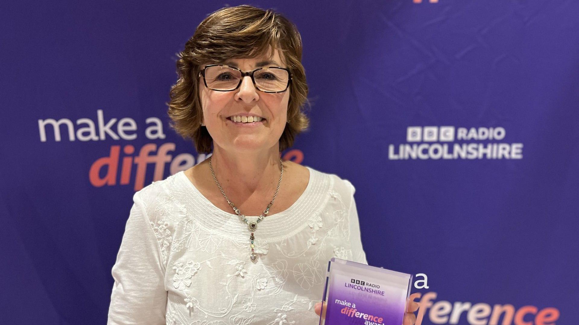 A woman smiles at the camera. She has a short brown haircut and is wearing dark square-framed glasses. She is wearing a white patterned top with a long silver necklace and is holding a glass trophy - with BBC Make A Difference written on it. She is smiling and behind her (although slightly blurred) is a purple backdrop with 'BBC Make A Difference' branding on it. 