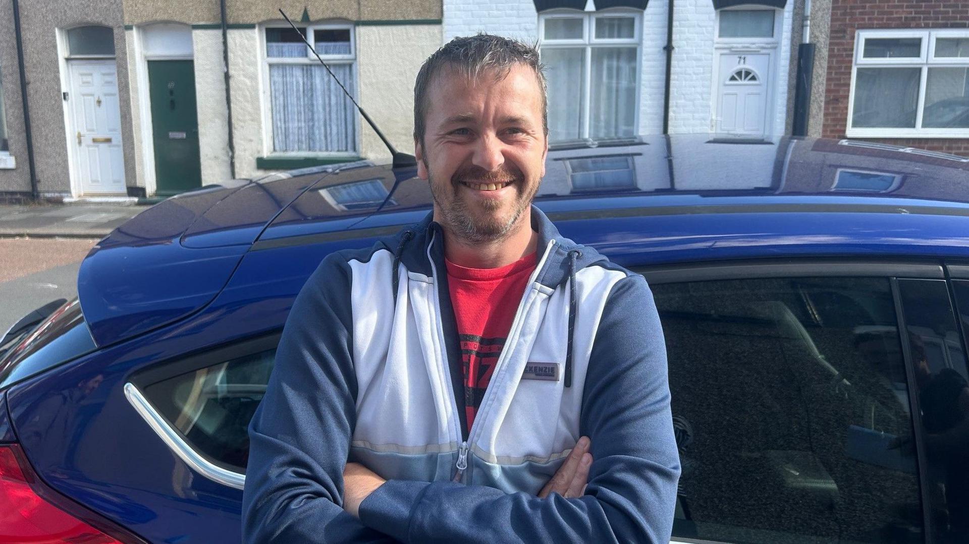 Scott Holliday stands outside a car with his arms folded. He is smiling. He has short grey hair and is wearing a grey and white hoodie.