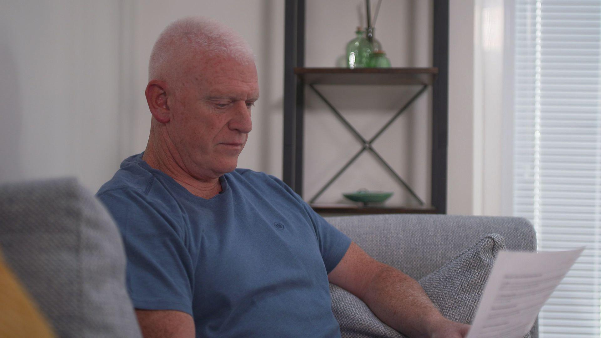Paul Harrison has short grey hair and is wearing a blue t-shirt. He is sitting on a grey sofa in his living room and reading a letter.