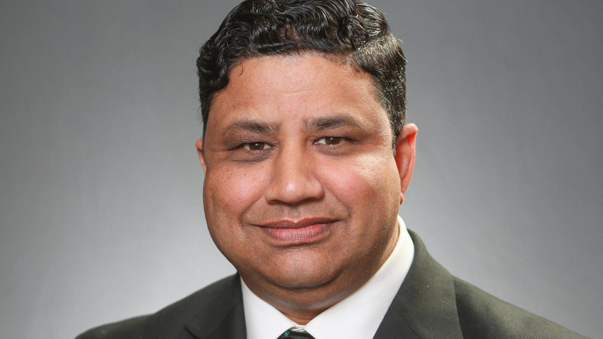 Naz Islam with short dark hair smiling at the camera and wearing a grey jacket and tie