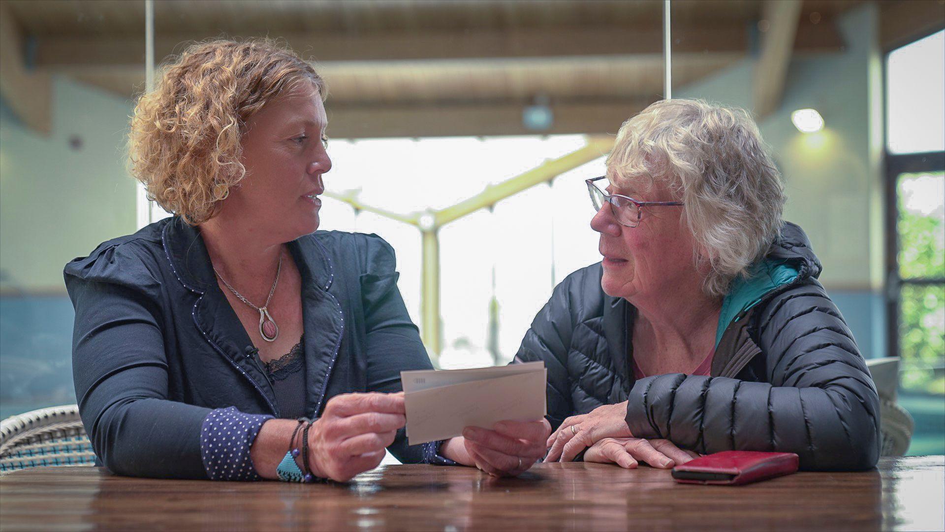 Two women sat at a table next to each other, looking at family photographs. They look at each other as they hold the photos. 
The woman on the left is wearing a navy jacket and has short ginger curly hair. The women on the right has short grey hair, glasses and is wearing a dark coat. 