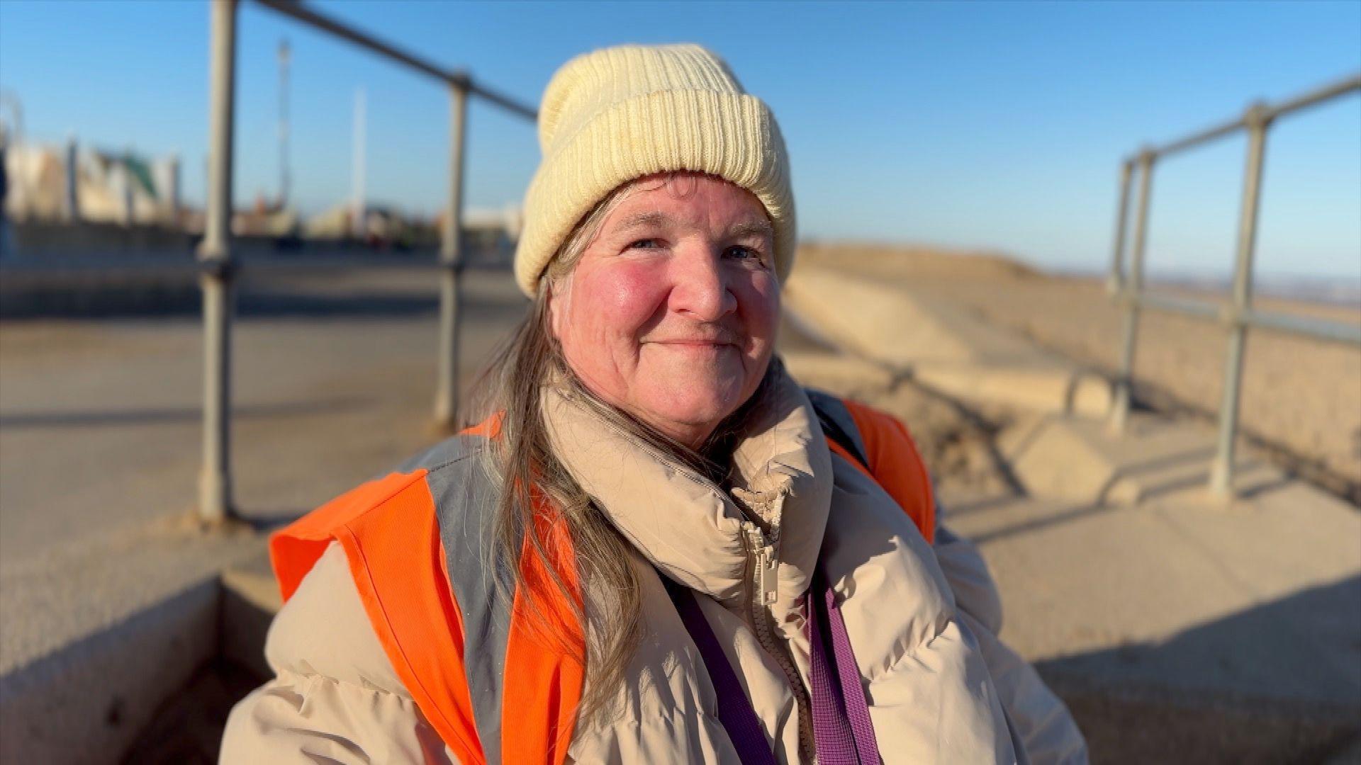 A woman with long hair wearing a cream winter beanie hat with a beige coat and orange high vis jacket. She is smiling into the camera with rosy cheeks and you can sea a beach in the background.