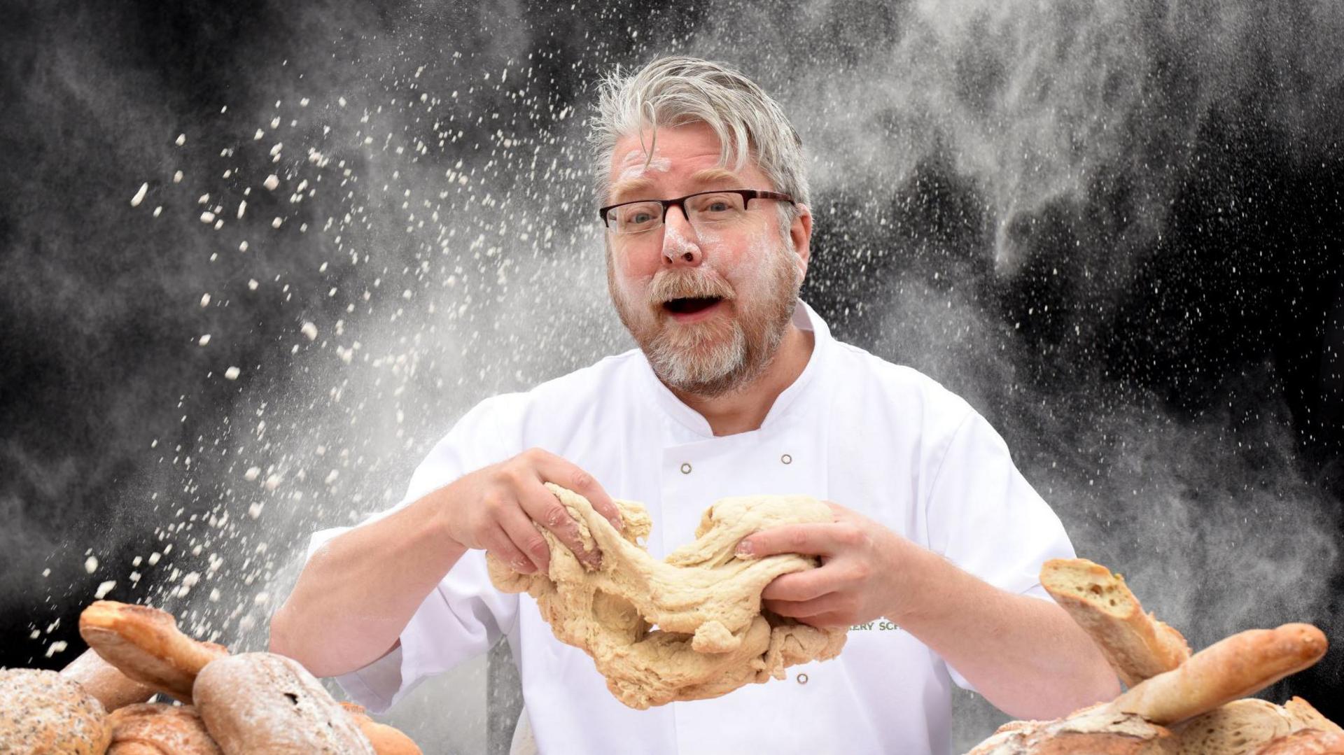 Richard Hughes in chef's whites, kneading dough and surrounded by loaves