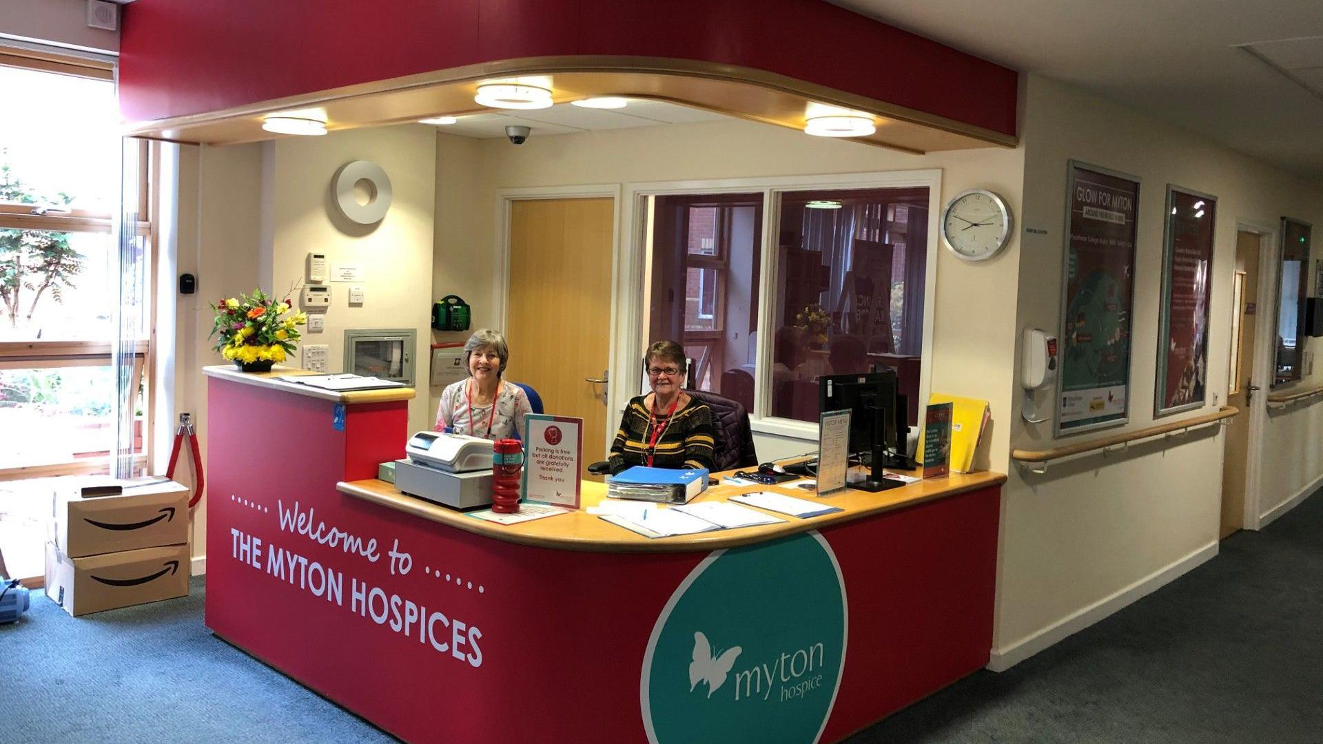 A reception area. It is decorated with a sign reading - welcome to The Myton Hospices. Two women are sat behind a desk and smiling to the camera. 