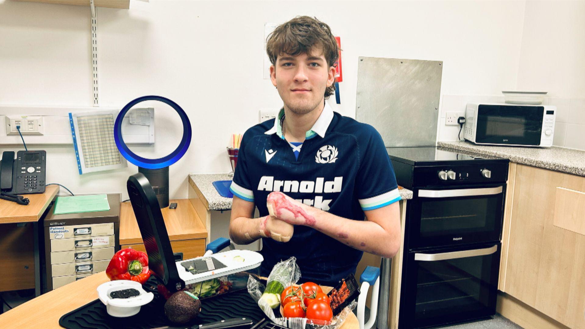 Hamish ( wearing a dark blue sports top) poses for a photo behind some food and cooking equipment. He has no hands.