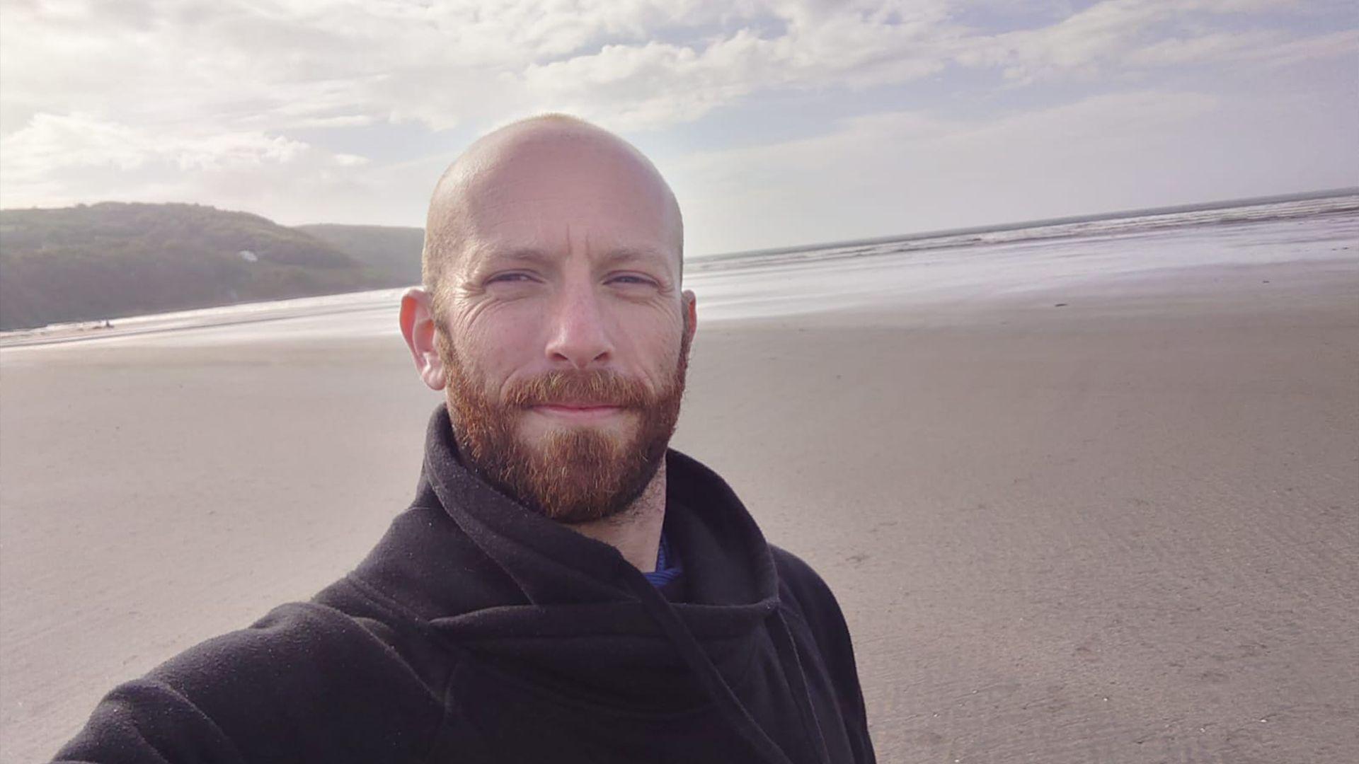 Dyn yn sefyll ar draeth ac yn edrych i'r camera, y môr yn y cenfdir