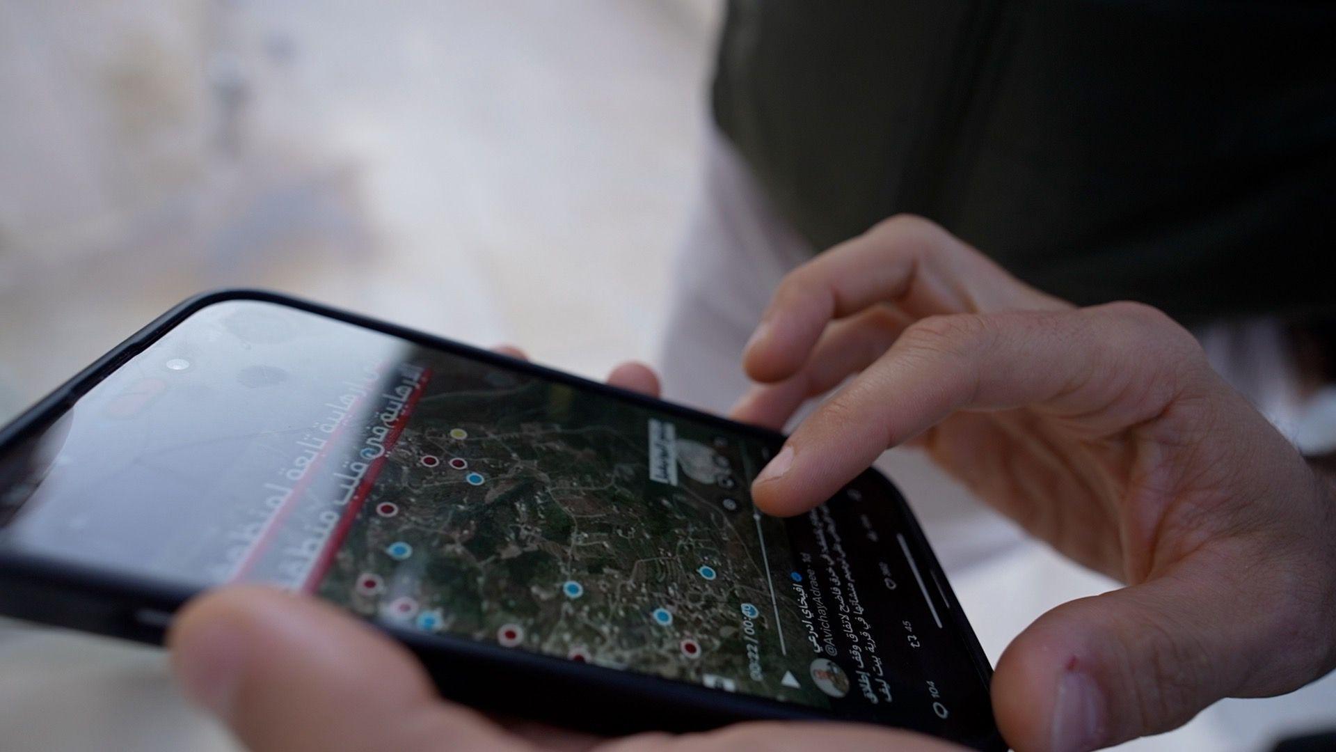 Hands hold a mobile phone, on which can be seen a satellite image with red and blue dots marking locations.