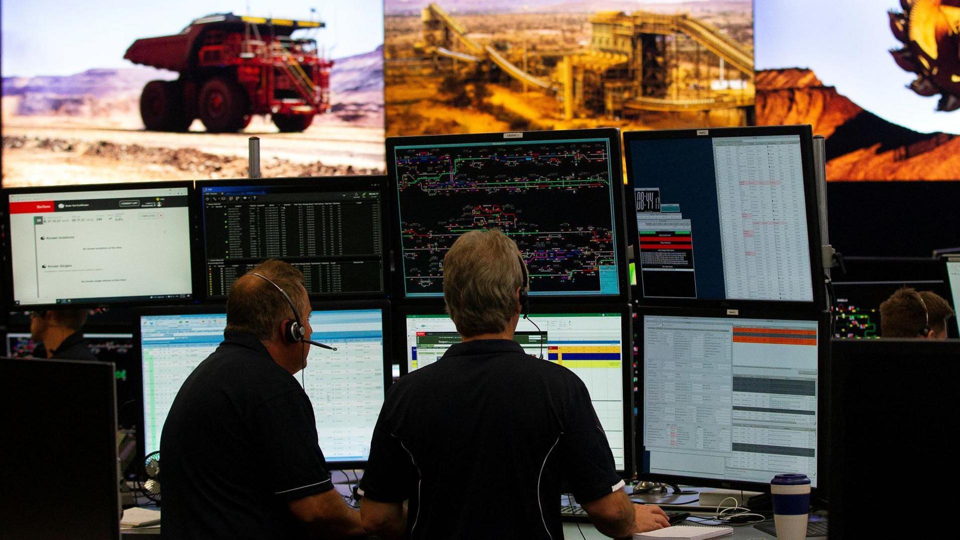 Two men sit at a desk with multiple screens monitoring trucks and other mining equipment