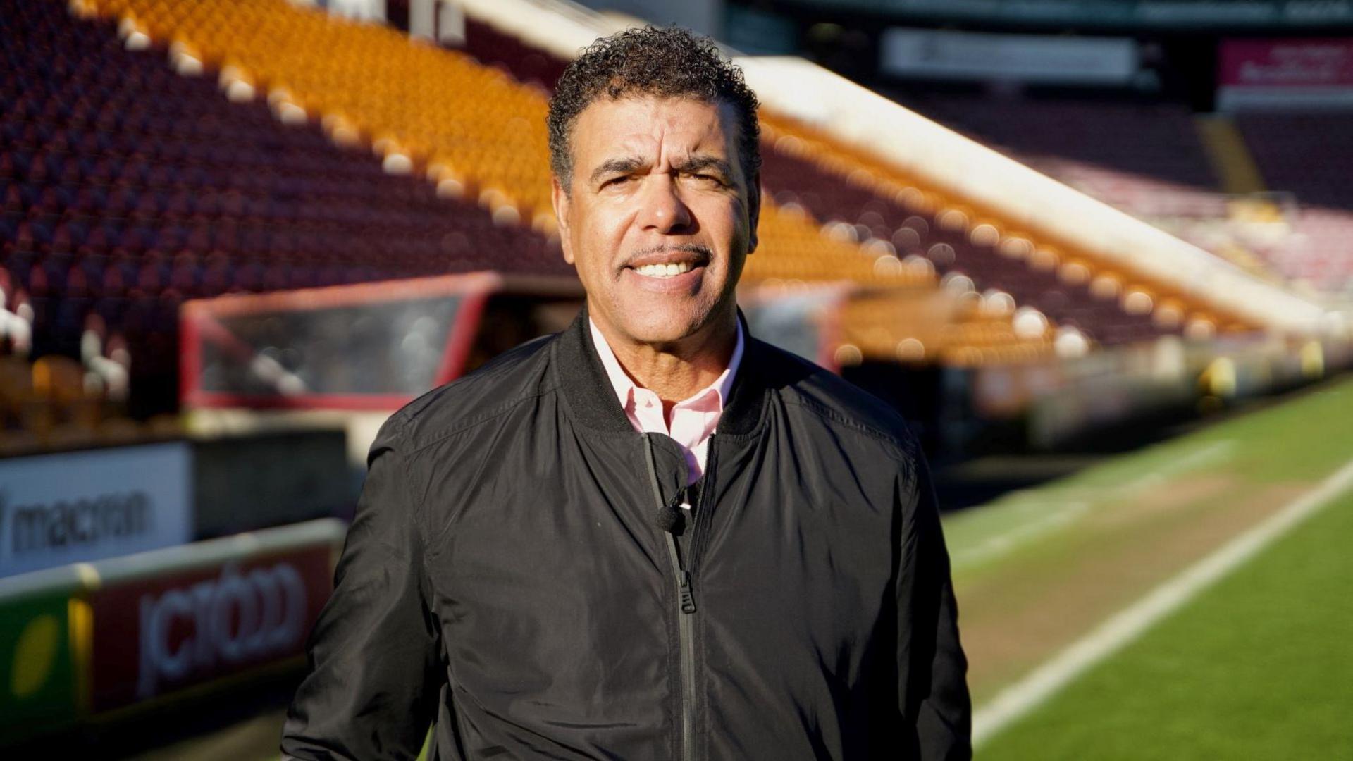 Chris Kamara stands on the pitch at Valley Parade. He is wearing a black jacket and white shirt.