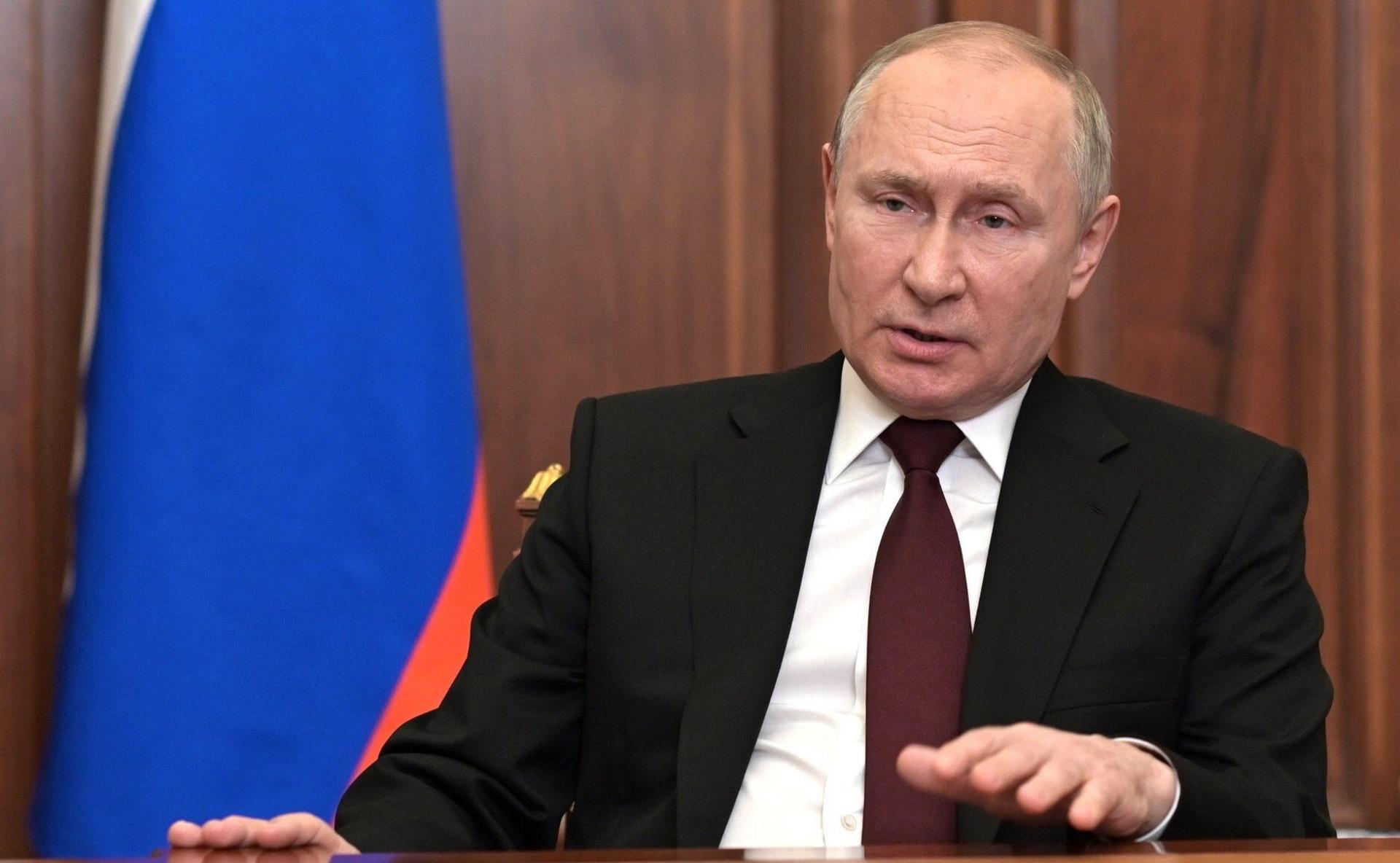 Vladimir Putin pictured delivering an TV address in Feb 2022 as he announced Russia's invasion into Ukraine. He is wearing a suit and is sitting in a wood-panelled room with a Russian flag to the left of the photo.