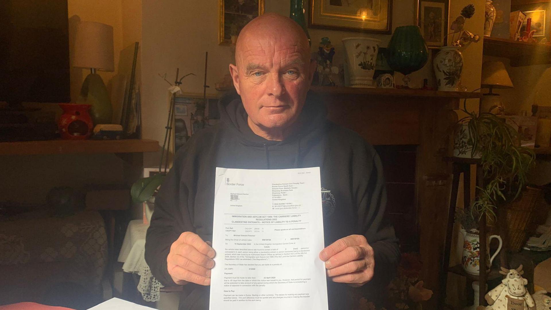 Michael Fletcher sitting in a dark room, holding up a letter which breaks down the details of his fine from Border Force, which he received in the post.