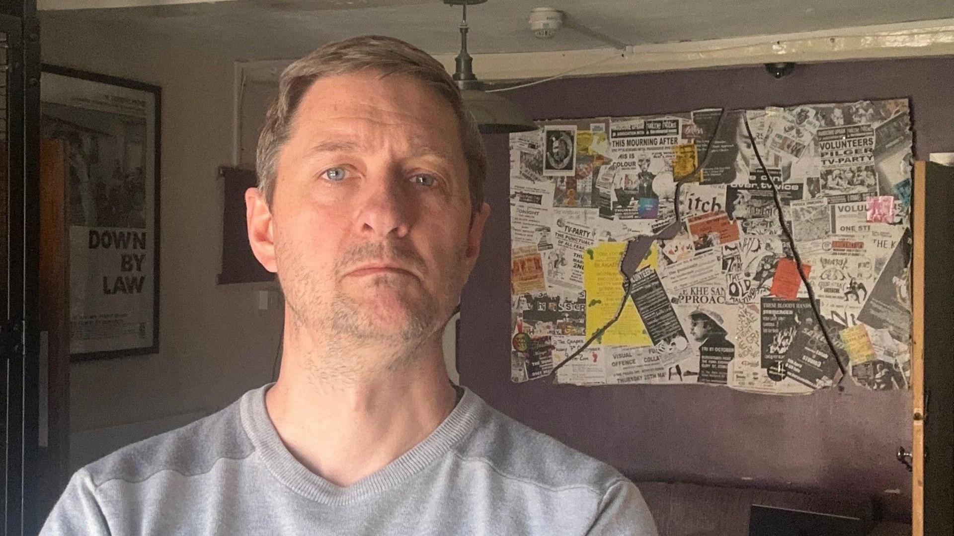 A head and shoulders image of Neil Attridge. He is wearing a grey jumper and looks glum. He is standing in the front of his recording studio.