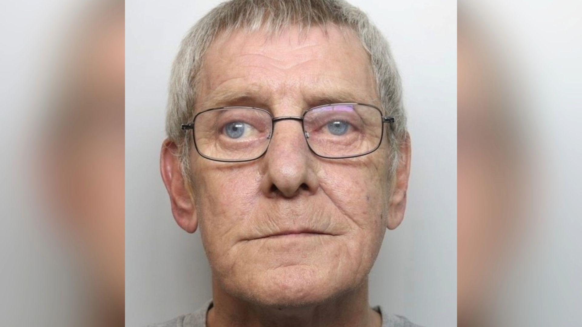 A mugshot of an elderly man with short grey hair and glasses. He is wearing a grey top.