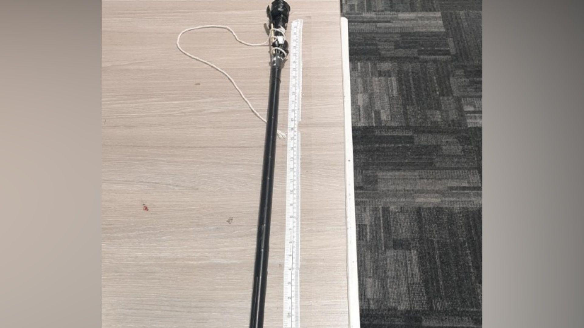 A black homemade blowpipe on a table alongside a measuring tape, viewed from above. There is a grey patterned carpet beneath the table.