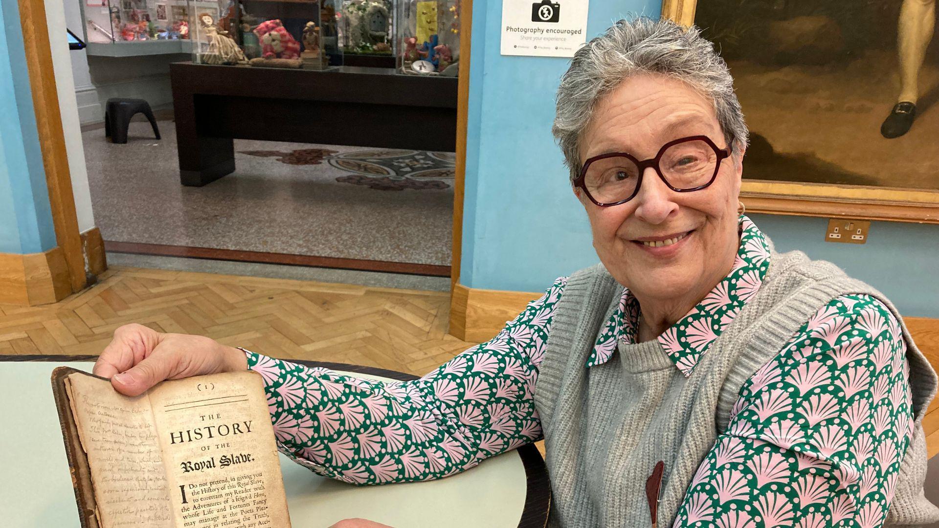 A woman with brown glasses and short grey hair. She is smiling and sitting down whilst holding a book open.