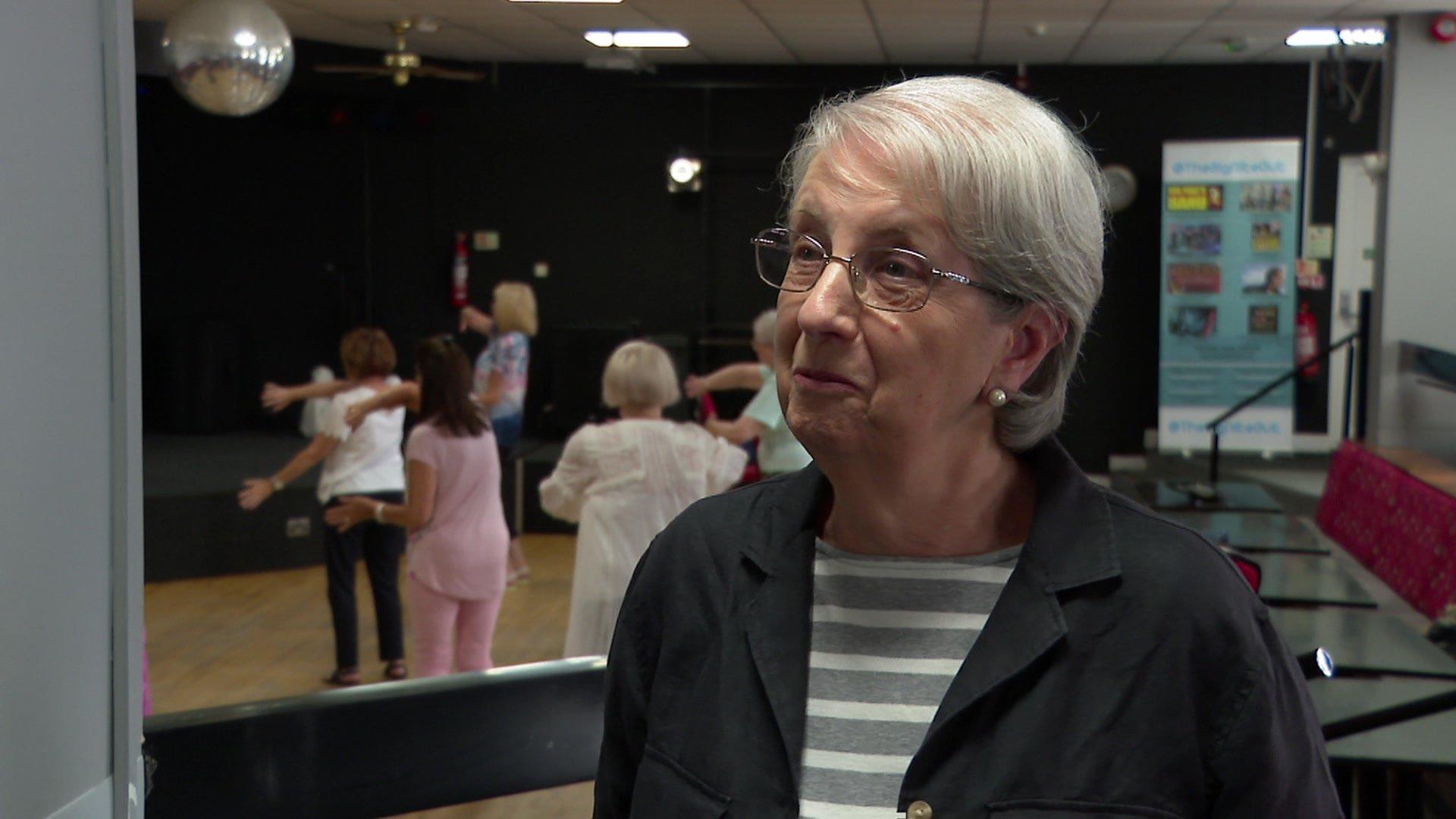 A lady with glasses in a hall. Behind her are a group of people taking part in a Tai Chi class