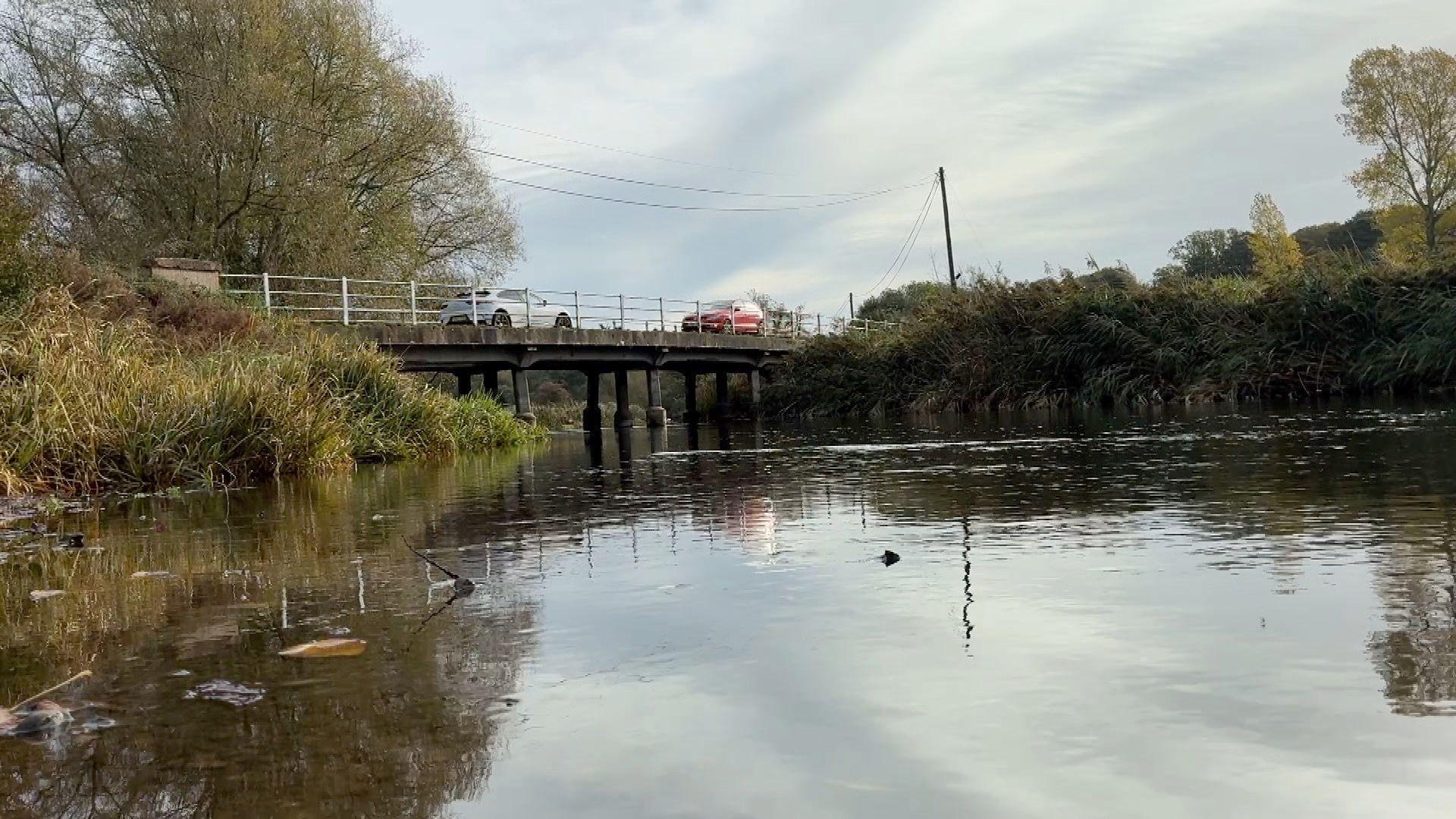 The photo is taken from water level, with the river in the foreground, with the light reflecting off its surface. There are grassy banks. In the background is the road bridge, with two cars crossing it going in different directions.