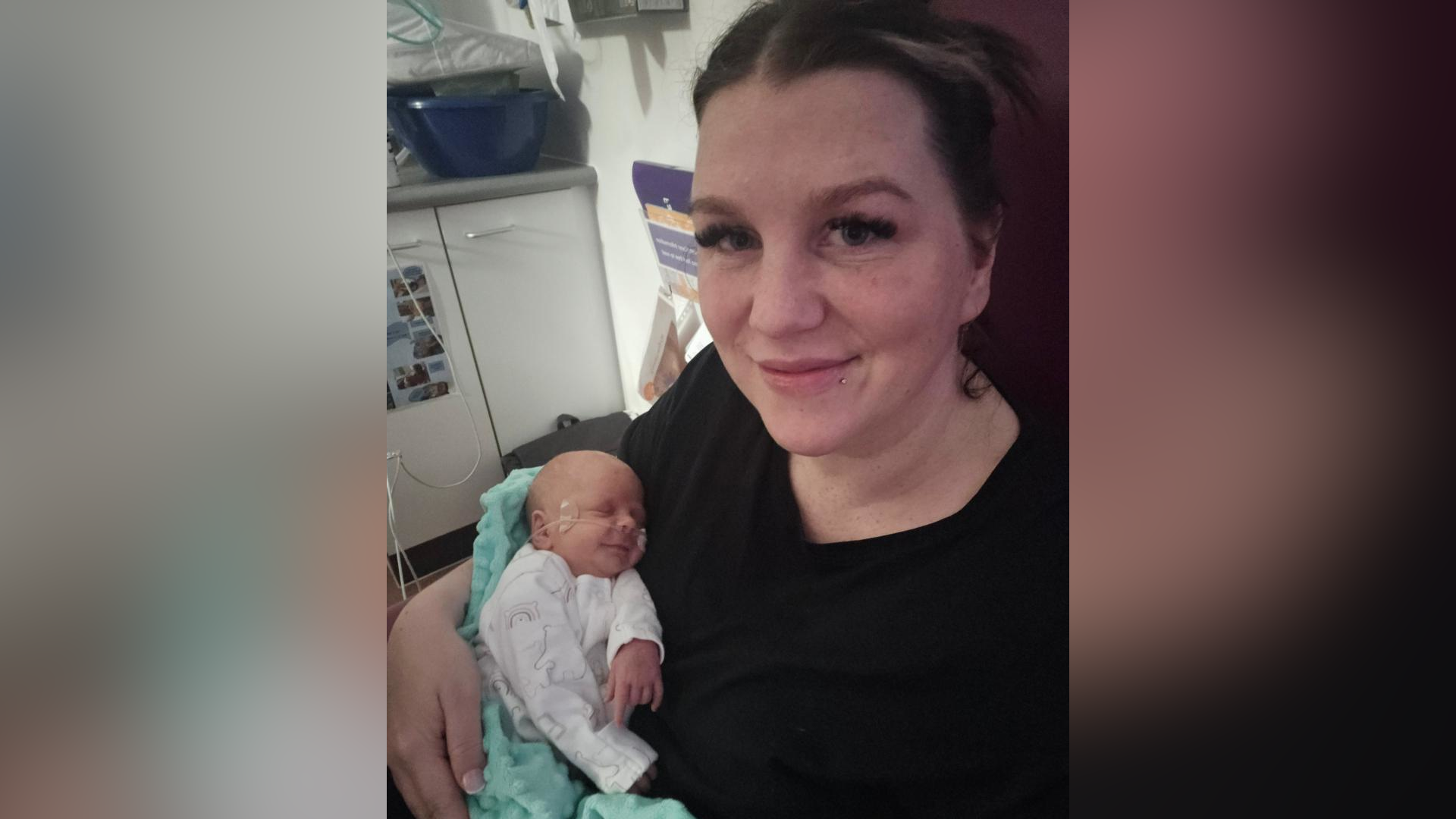 Ms Lazinska is smiling at the camera while in hospital, She is wearing a black top, has dark hair tied back and long eyelashes. She is holding her baby in her arms. He is very small and has a tube running under his nose. He is wearing a white baby grow and is wrapped in a blue blanket.