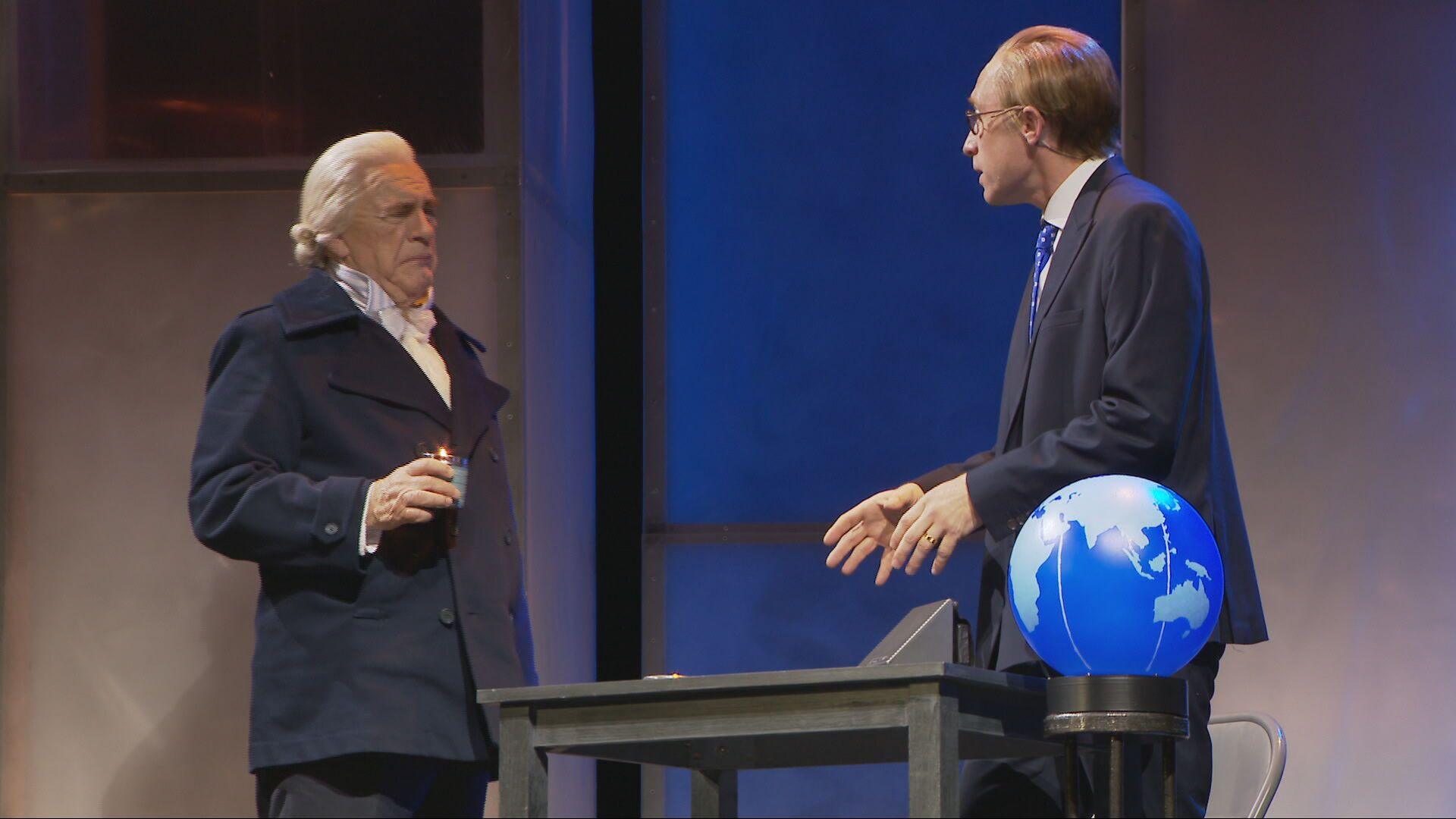 Brian Cox on stage portraying Adam Smith. He is wearing a blond/white wig, a dark jacket and a ruffled shirt. He is talking to a man in a suit and glasses and there is a table and globe on the stage  
