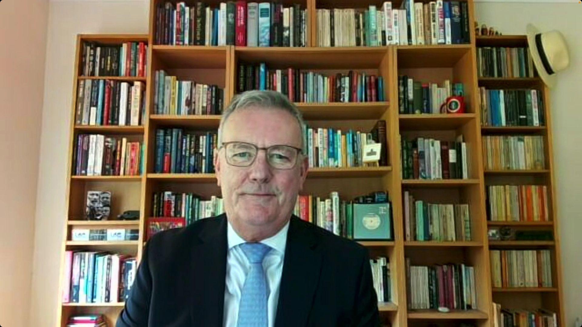 Screenshot of Ulster Unionist leader Mike Nesbitt on a zoom call wearing a black jacket, white shirt and light blue tie. He is sitting in front of a book case filled with books and pictures