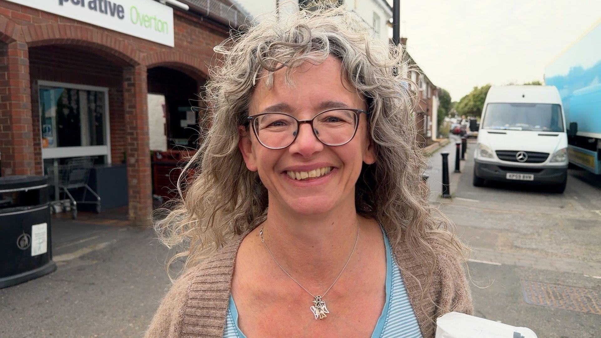 A woman with long, wavy grey hair, glasses, a grey cardigan and a light blue stripy top underneath, standing in front of a co-op shop and smiling.