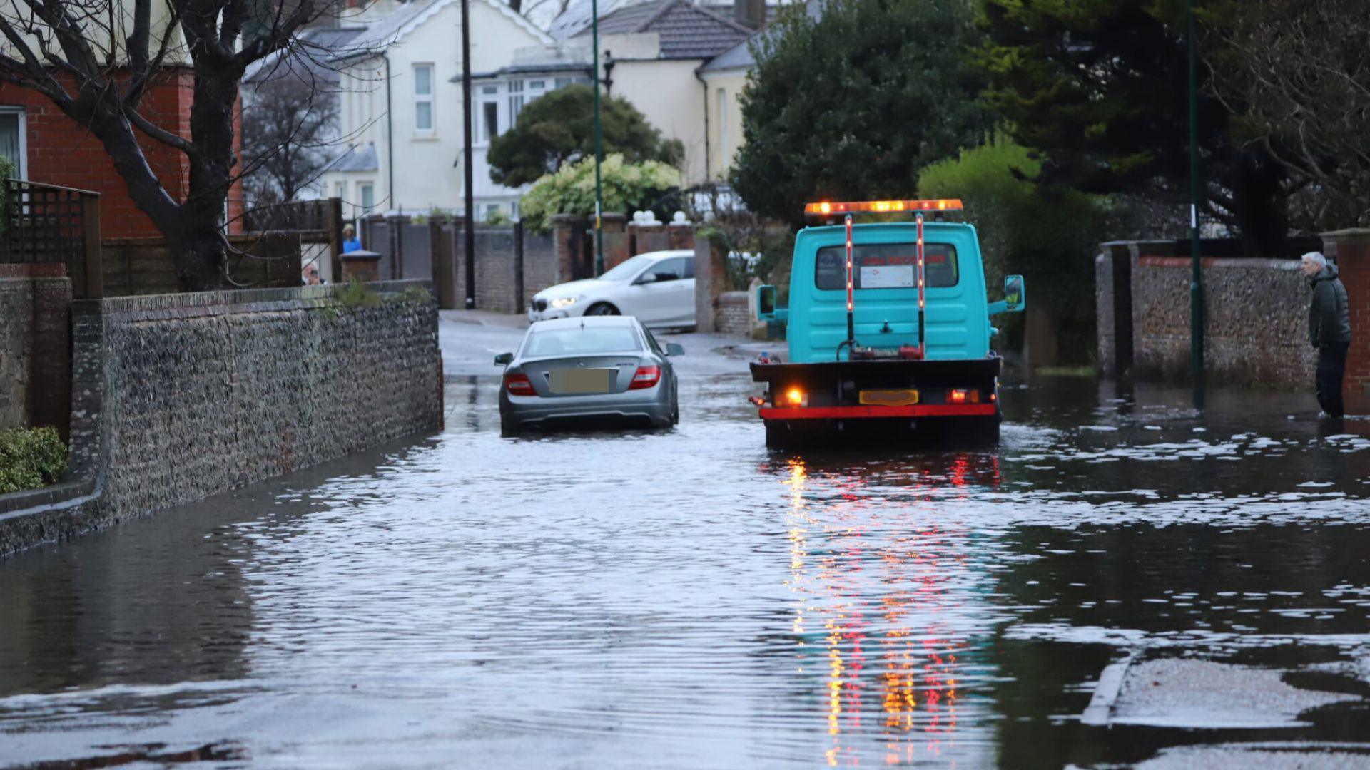 A silver car driving through deep water in a residential street. There is a blue truck to the right with orange flashing lights