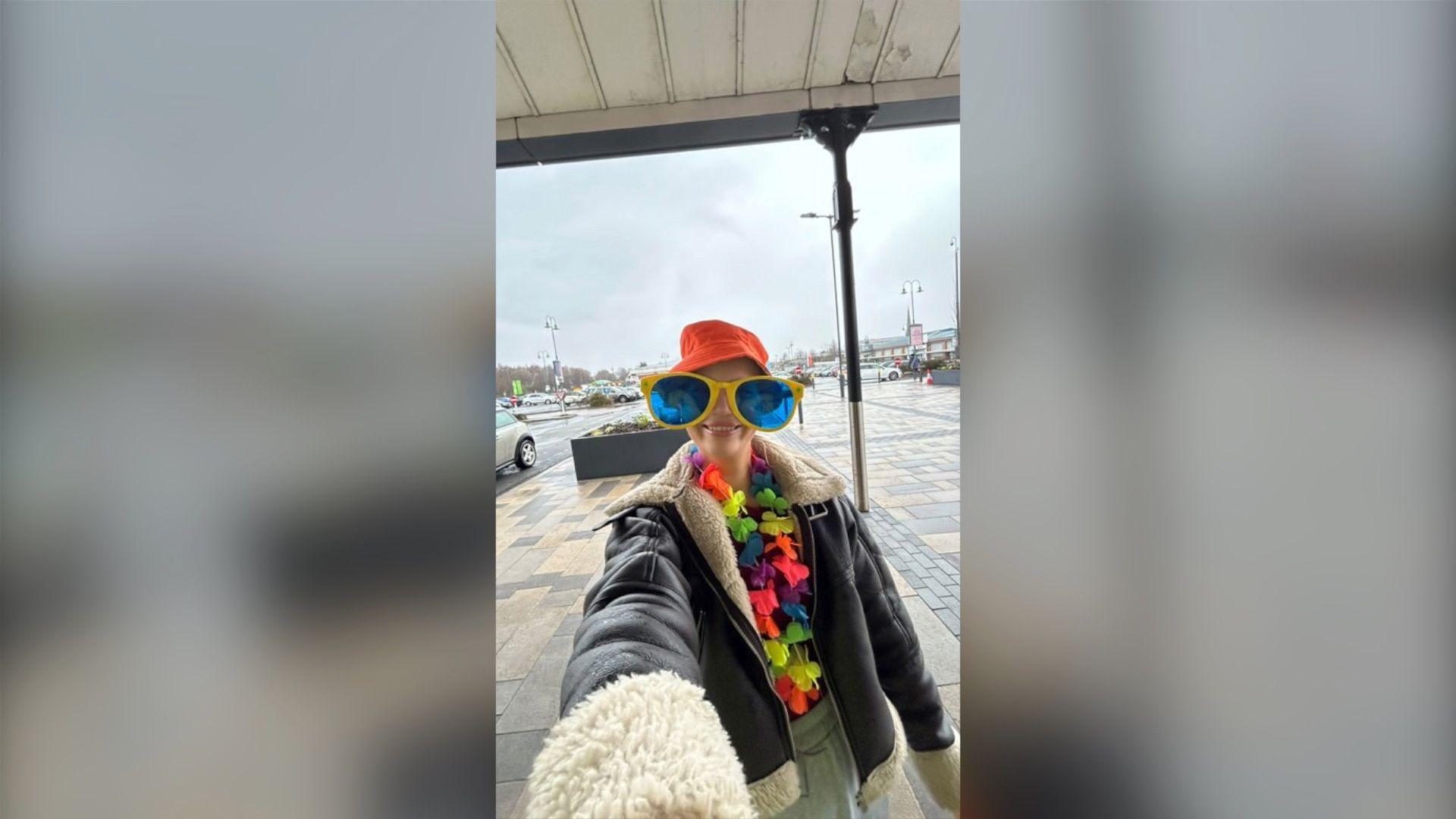 A woman is wearing oversized, comedy, yellow glasses, a rainbow flower garland round her neck and a bright orange bucket hat. She is smiling at the camera. She is wearing a leather jacket.