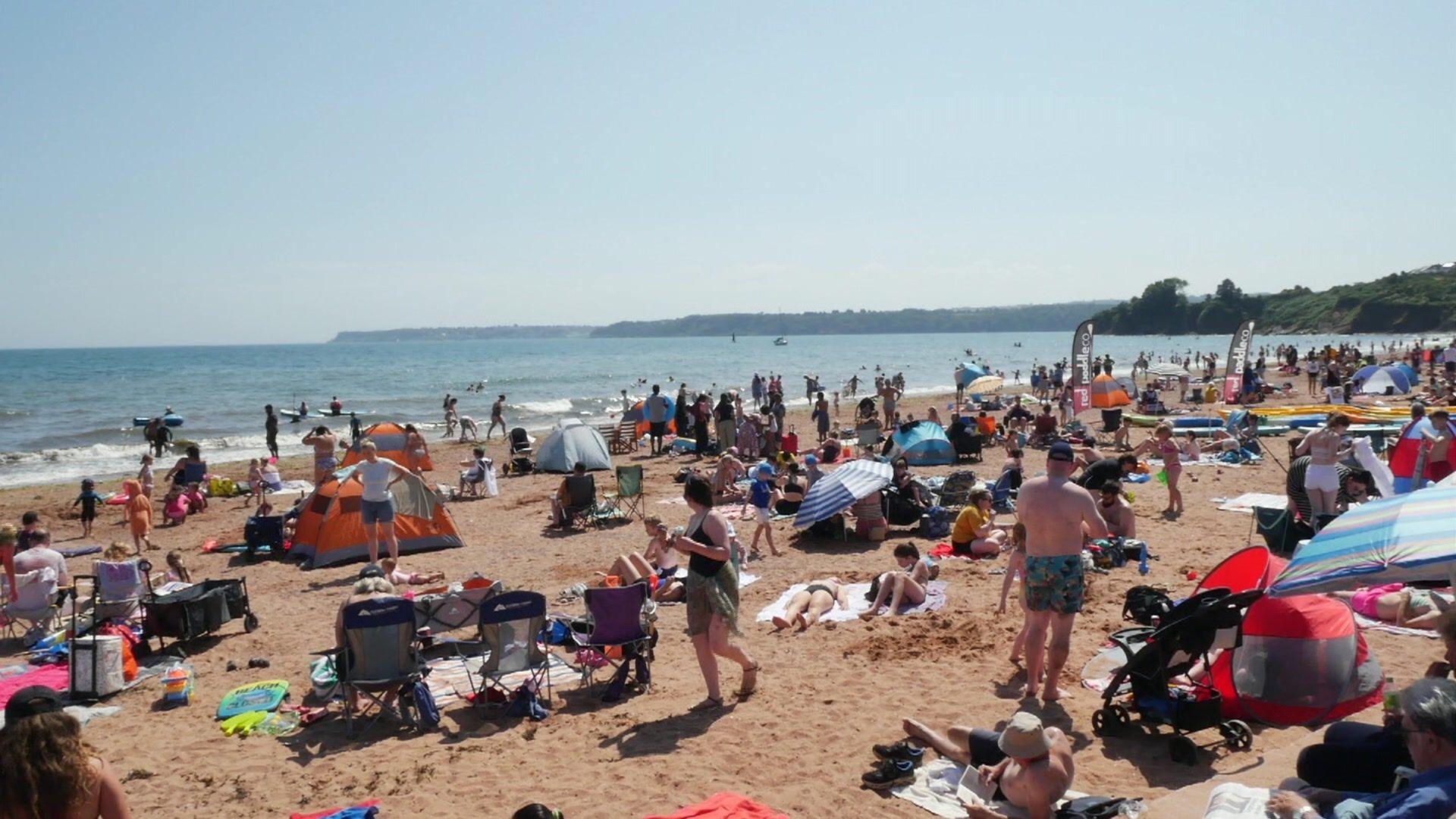 A photo which shows Goodrington Sands in Paignton. Hundreds of people are on the sand with deck chairs, towels and tents, as well as umbrellas. Some people are sunbathing