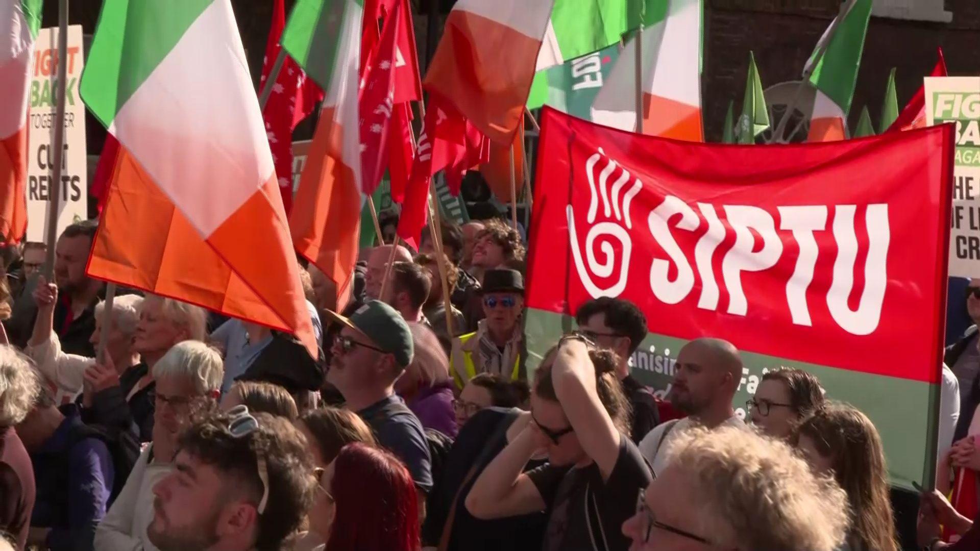 A crowd of people have gathered in the street. Some are waving Irish flags, while others are holding flags belonging to several different unions.