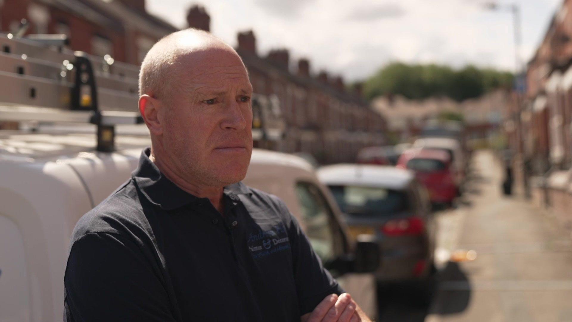 Andrew Hay is looking to the right of the camera. He is bald, is wearing a dark T-shirt, and has his arms folded.