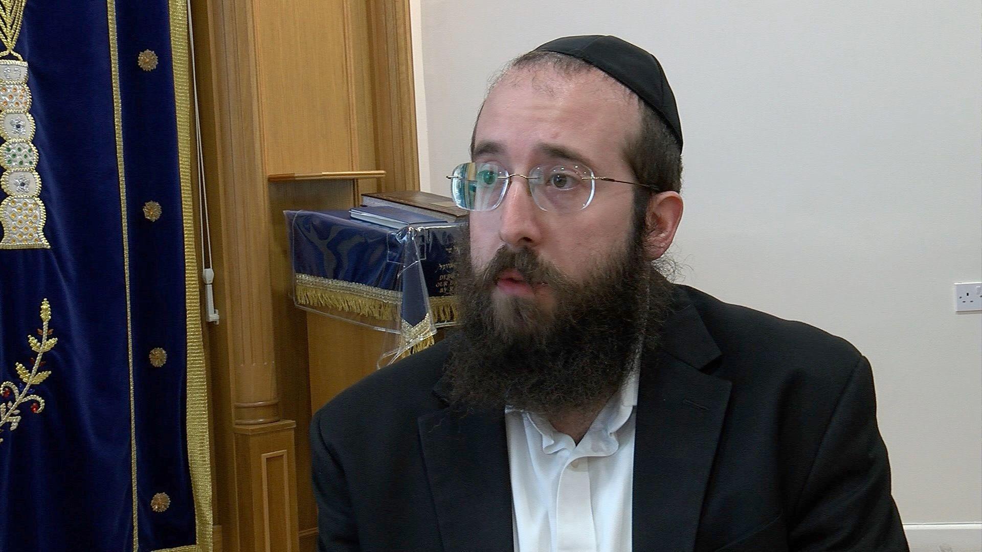 Rabbi Bentzion Alperowitz, looking left out of frame, wearing glasses and a yarmulke or traditional Jewish skullcap. He has a long beard and is wearing a black blazer and white shirt.