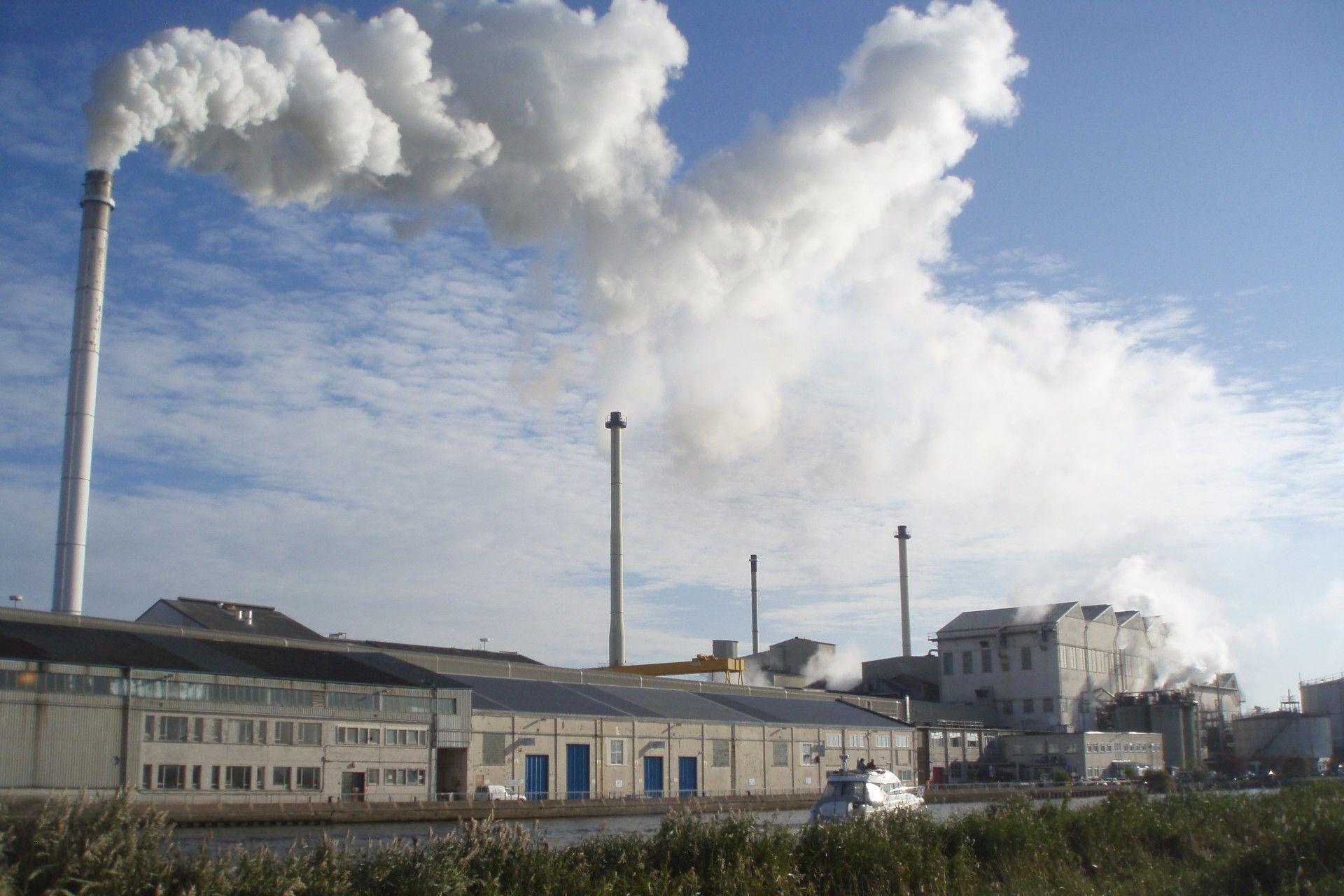 A factory site with a large metal cylinder and buildings can be seen from the other side of a river, with clouds of steam rising into the air on a sunny day.
