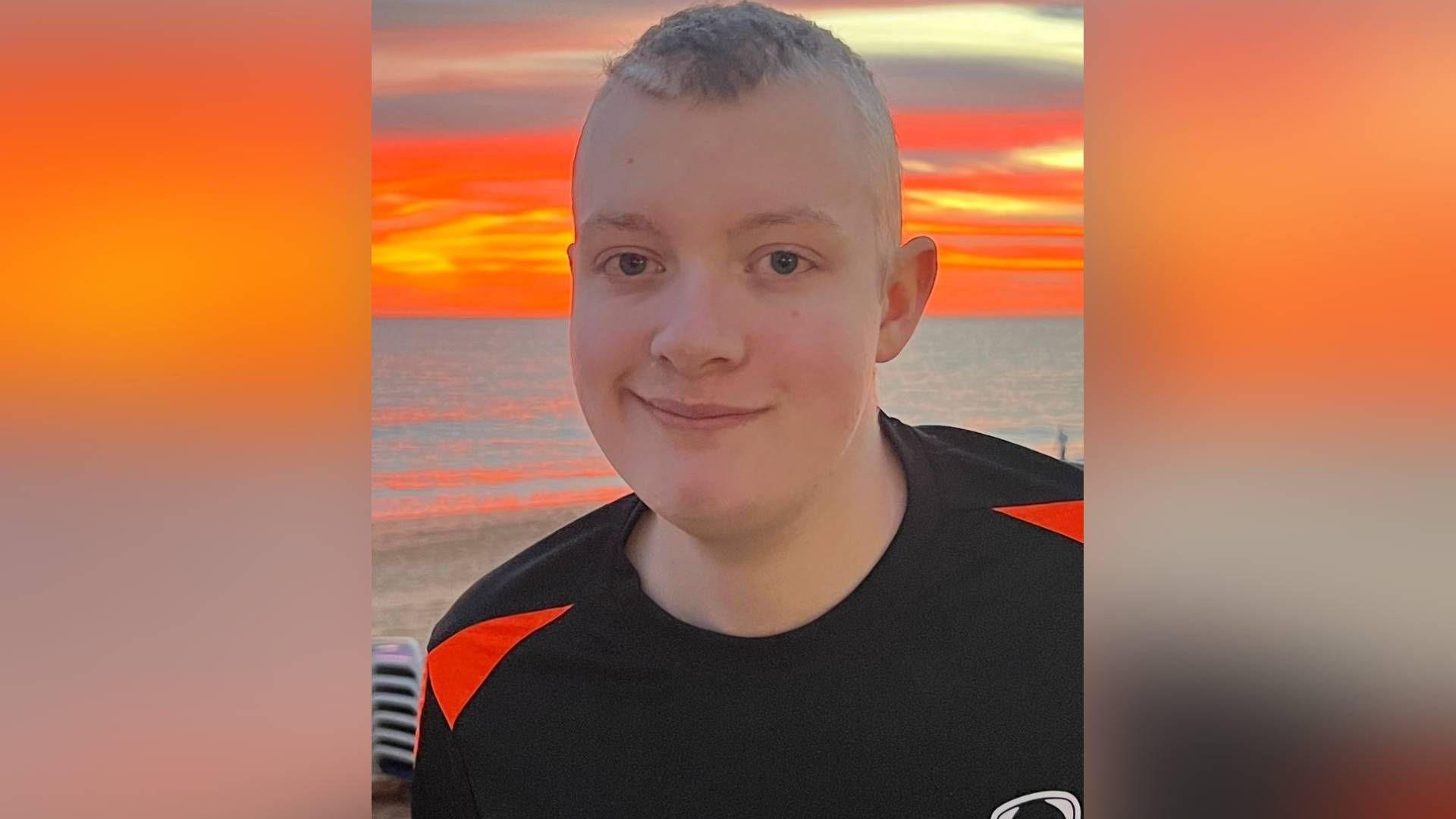 Aiden standing in front of a vibrant orange sun setting beyond the sea. He wears a black football top and smiles at the camera. He has blue eyes with short cropped lighter hair. 