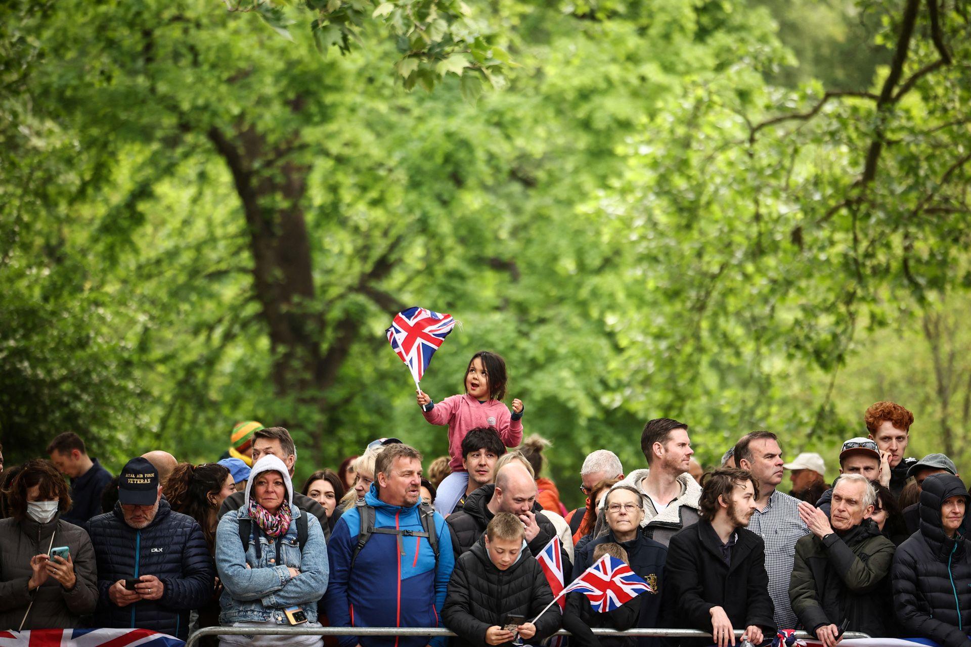 A small girl wearing a pink jumper waves a Union Jack in her hand while on the shoulders of a member of the crowd lining the Mall, with other members of the public around them and green foliage in the background.