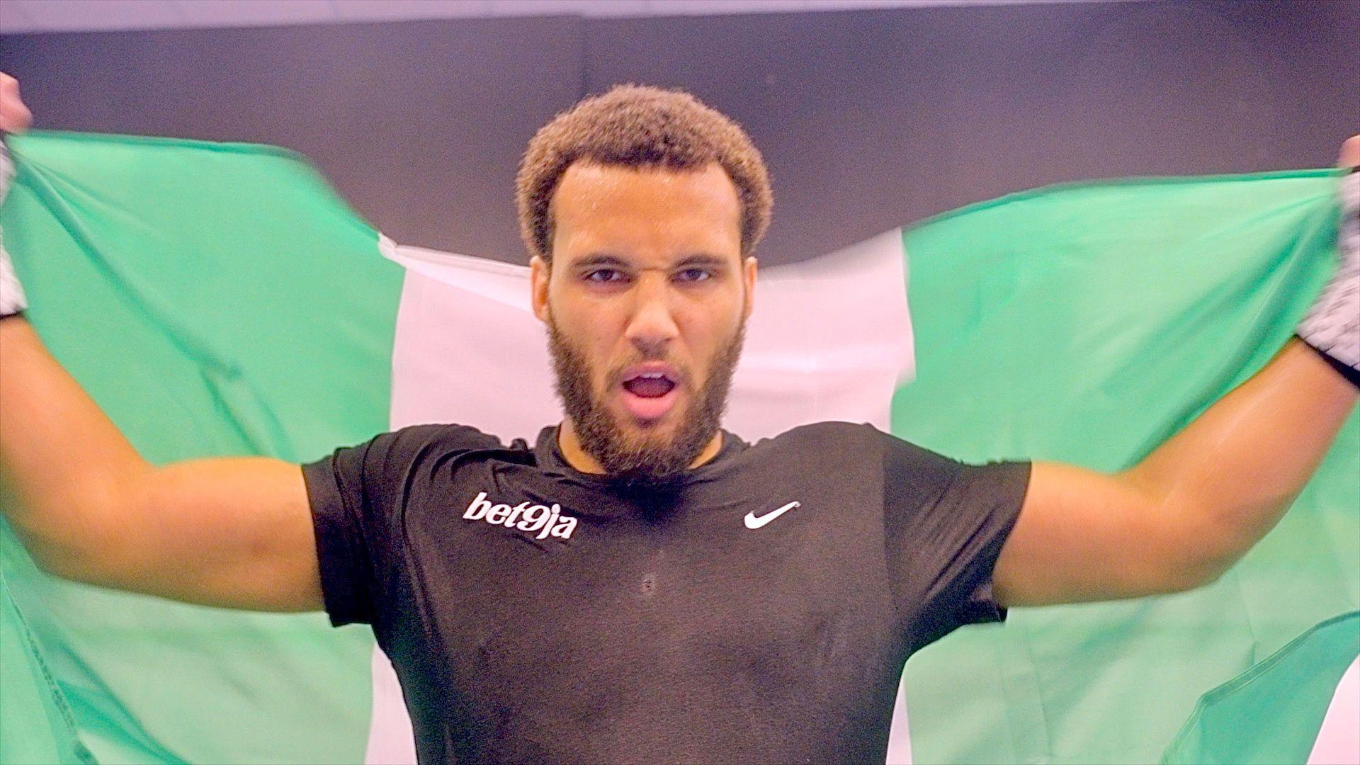 Moses Itauma visible from stomach up wearing a black short-sleeved training top extends his arms out wide to hold up a Nigerian flag behind himself. He is looking down into the camera with a fierce expression on his face and his mouth slightly opened in what looks like a roar