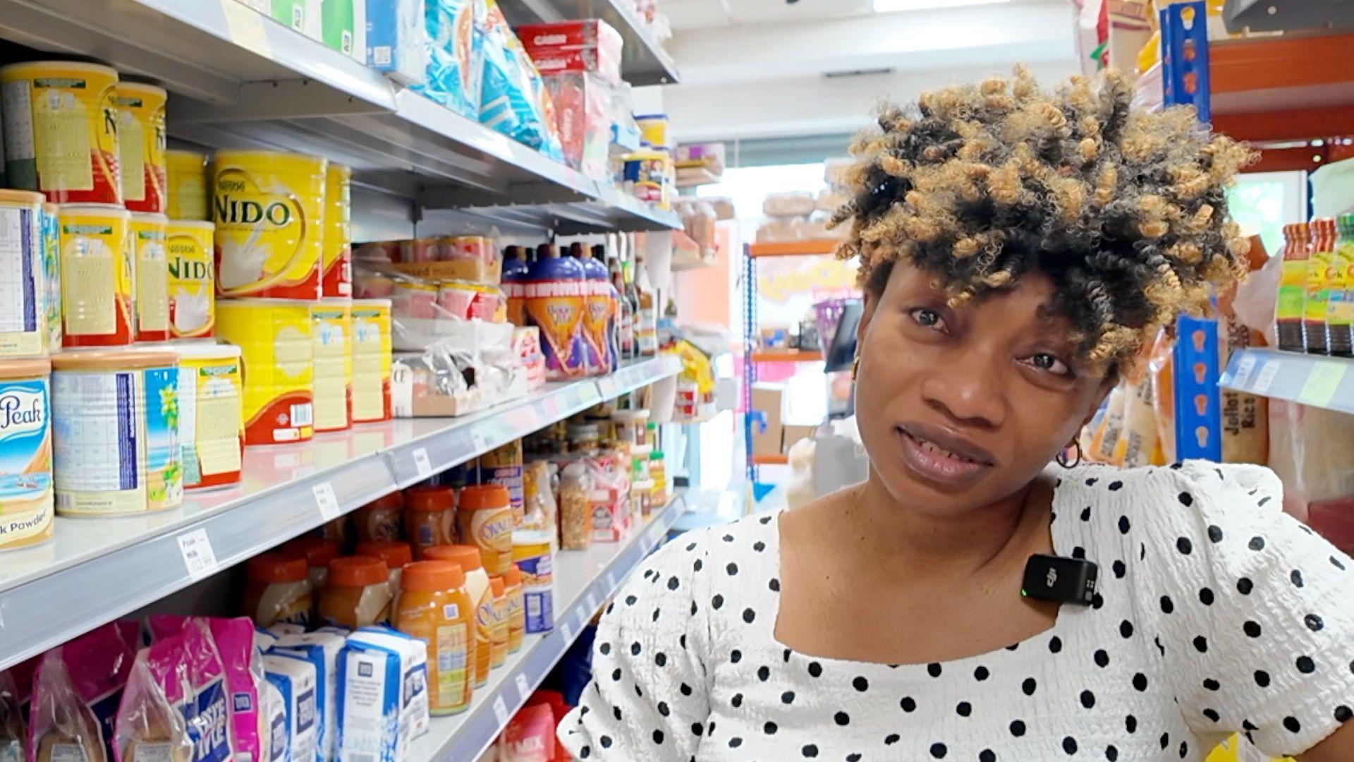 Nnenna Okonkwo tilts her head as she looks at the camera with tears in her eyes. She is standing in an aisle of a shop with food on the shelves beside her.