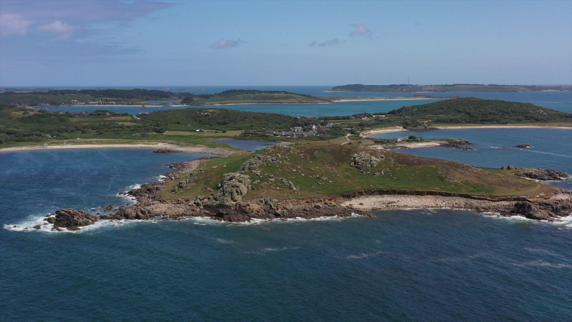 The island of Bryher is shown with rocky hills, sandy beaches and bodies of water beneath a blue sky scattered with clouds.