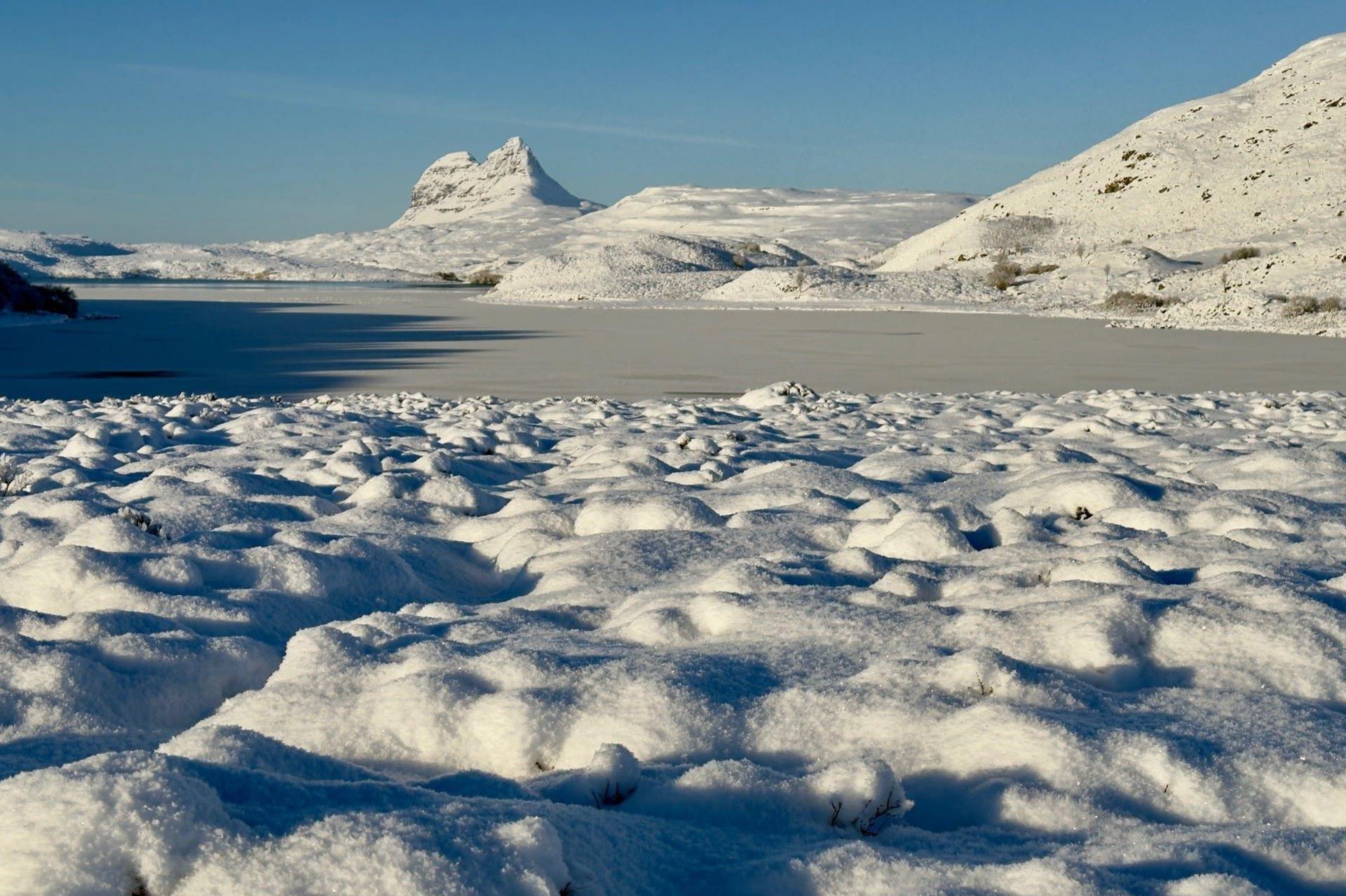 The ground is blanketed by thick snow and ice covers a loch in the Highlands in the distance is snow-covered Suilven.