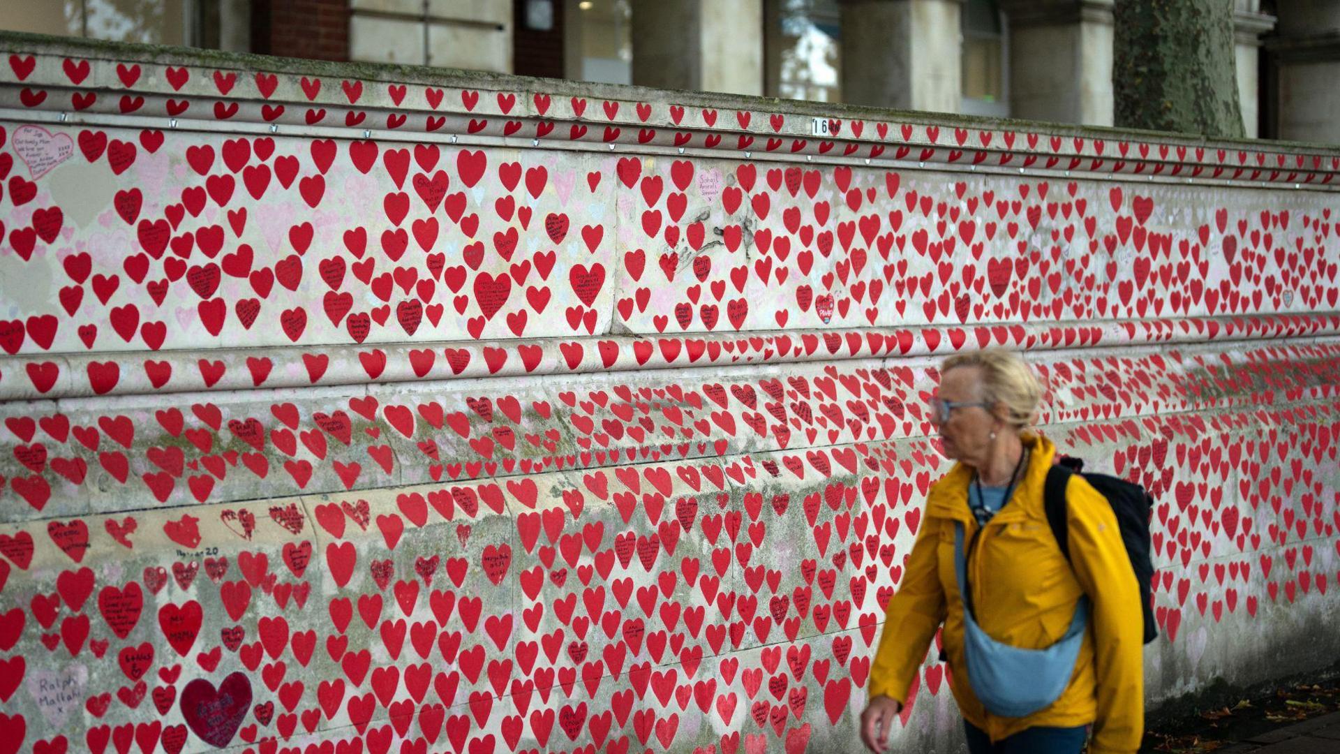  A woman wearing a yellow coat walks past the Covid memorial wall in London. It is a grey wall covered in red hearts