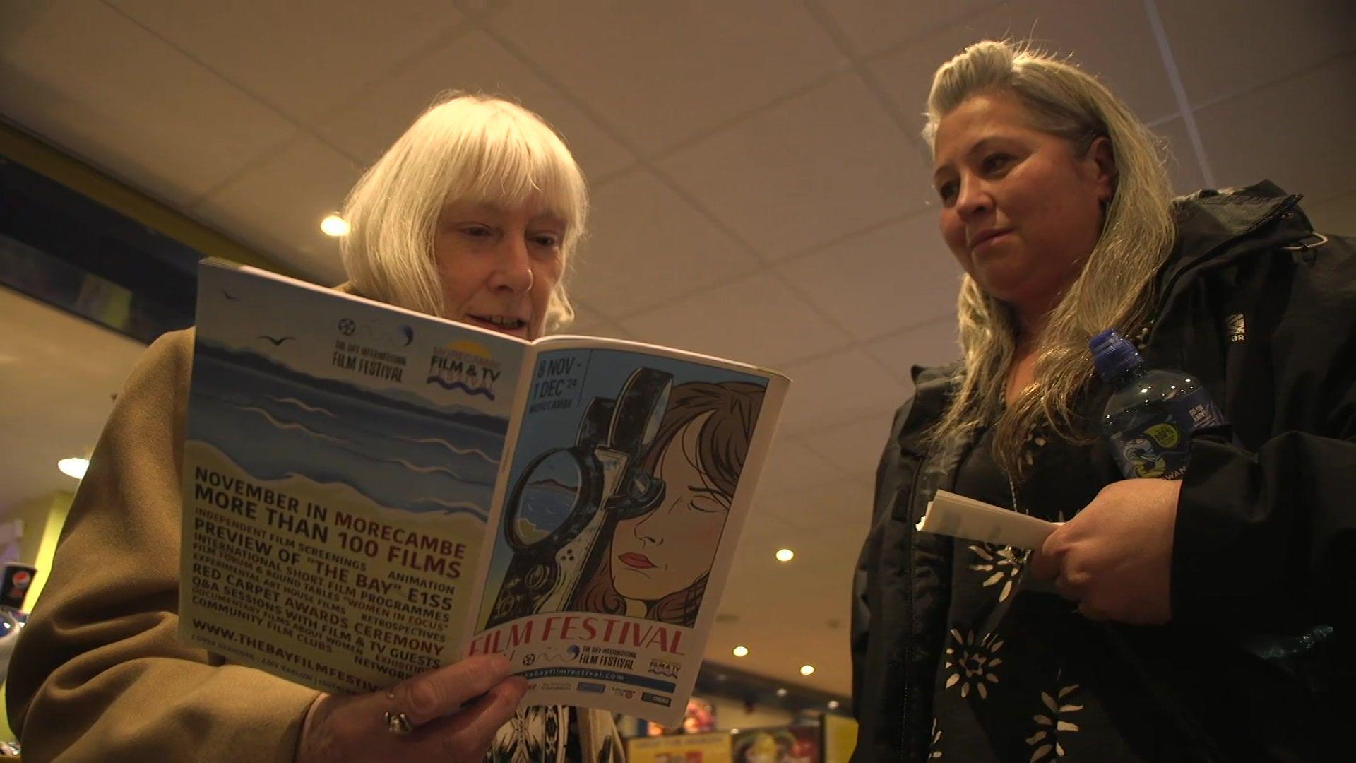 Two women are in the foyer of a cinema looking at a festival programme. One woman has short grey hair and is wearing a light coat and holding the programme. The second woman had long blonde hair and is wearing a black coat and is holding two cinema tickets.