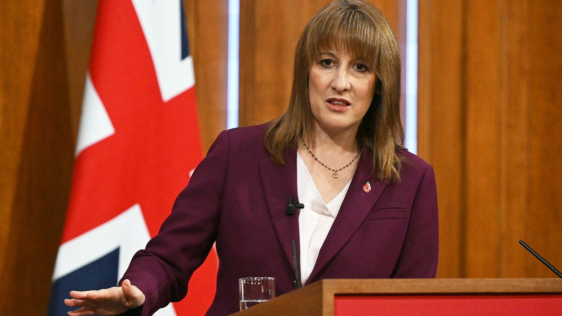 Rachel Reeves wearing a dark maroon jacket and white shirt, standing at a podium in front of a wood-panelled wall and a large Union Jack flag. She is gesturing with her right hand.