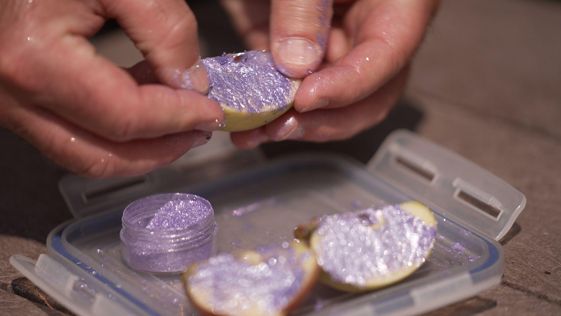 A man's hands holds an apple which is covered in purple glitter. Underneath the apple is a small glitter container with its cap removed.